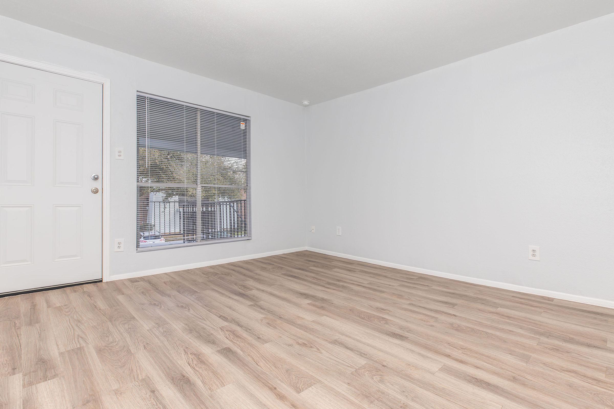 A spacious, empty room with light gray walls and a light wood floor. There is a large window with blinds on the left, letting in natural light. The front door is visible on the left side of the image, adding an inviting touch to the interior space.