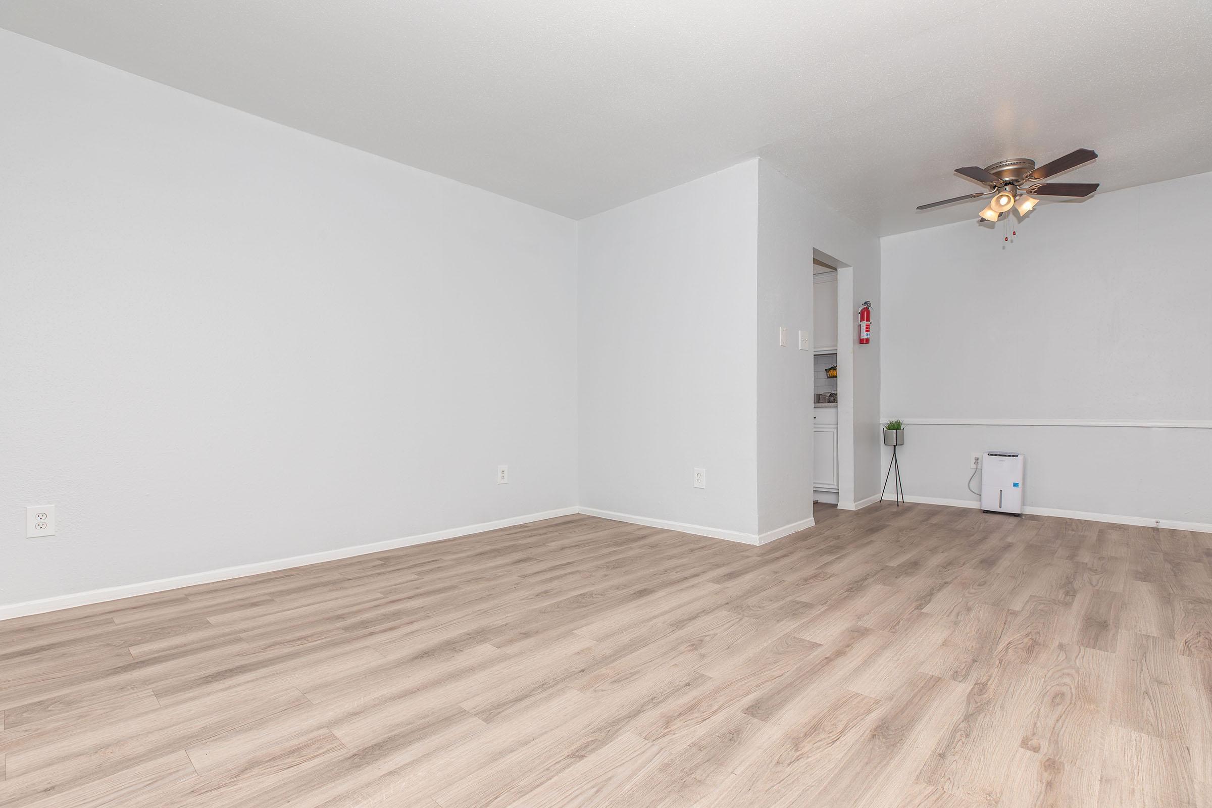 Empty room with light gray walls and modern wood-like flooring. A ceiling fan is visible, and there's a small area with a fire extinguisher on a wall. To the left, an open doorway leads to another space, suggesting a kitchen or utility area. Overall, the space is bright and uncluttered.