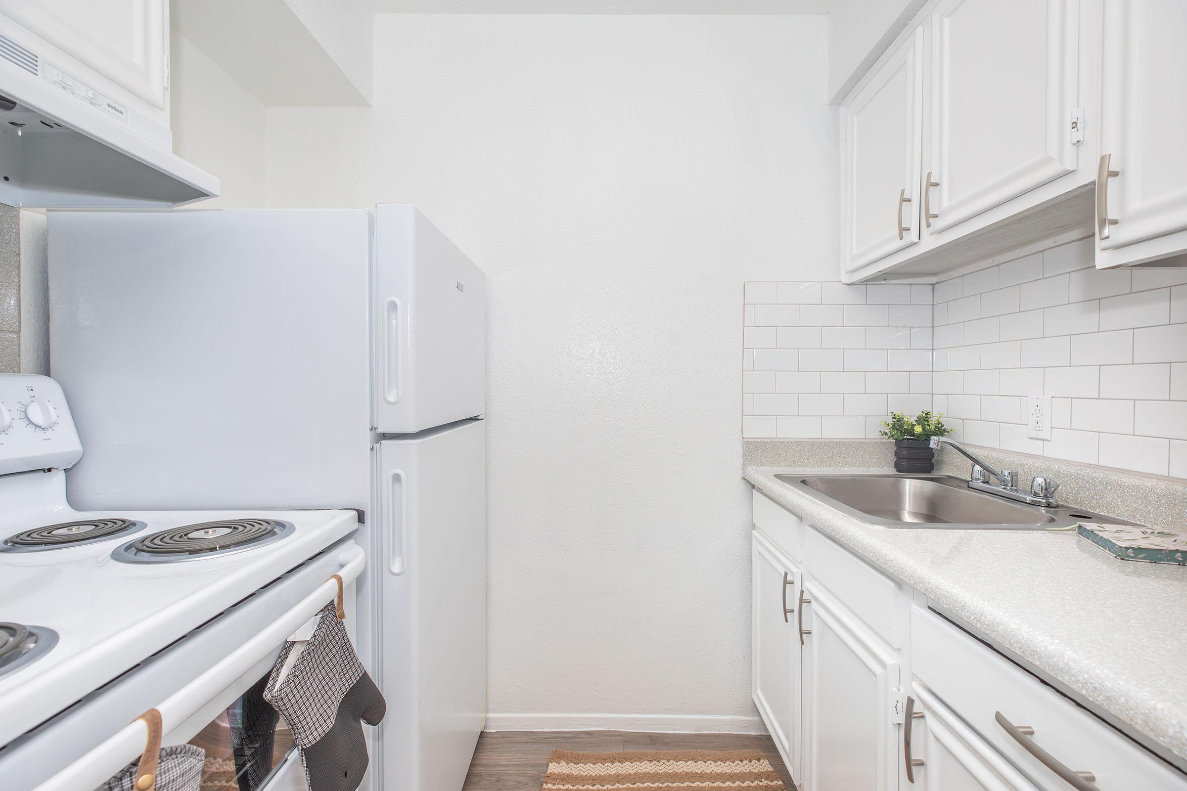Bright and clean kitchen featuring white cabinetry, a refrigerator, and a stove with an oven. The countertop has a small sink and a decorative plant. Subway tile backsplash adds a modern touch, with a neutral color scheme and warm wood flooring. A rug is placed in front of the sink for added comfort.