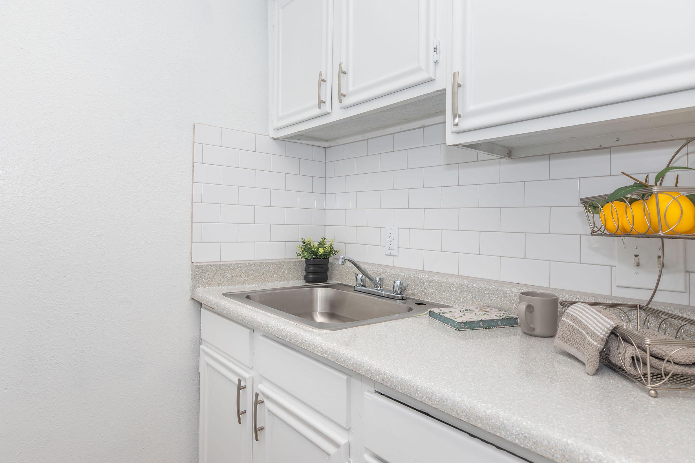 A clean, modern kitchen with white cabinets, a stainless steel sink, and a light-colored countertop. The backsplash features white tiles. A small potted plant sits on the counter, alongside a bowl of oranges and a stack of kitchen towels. A coffee mug is also visible on the countertop.