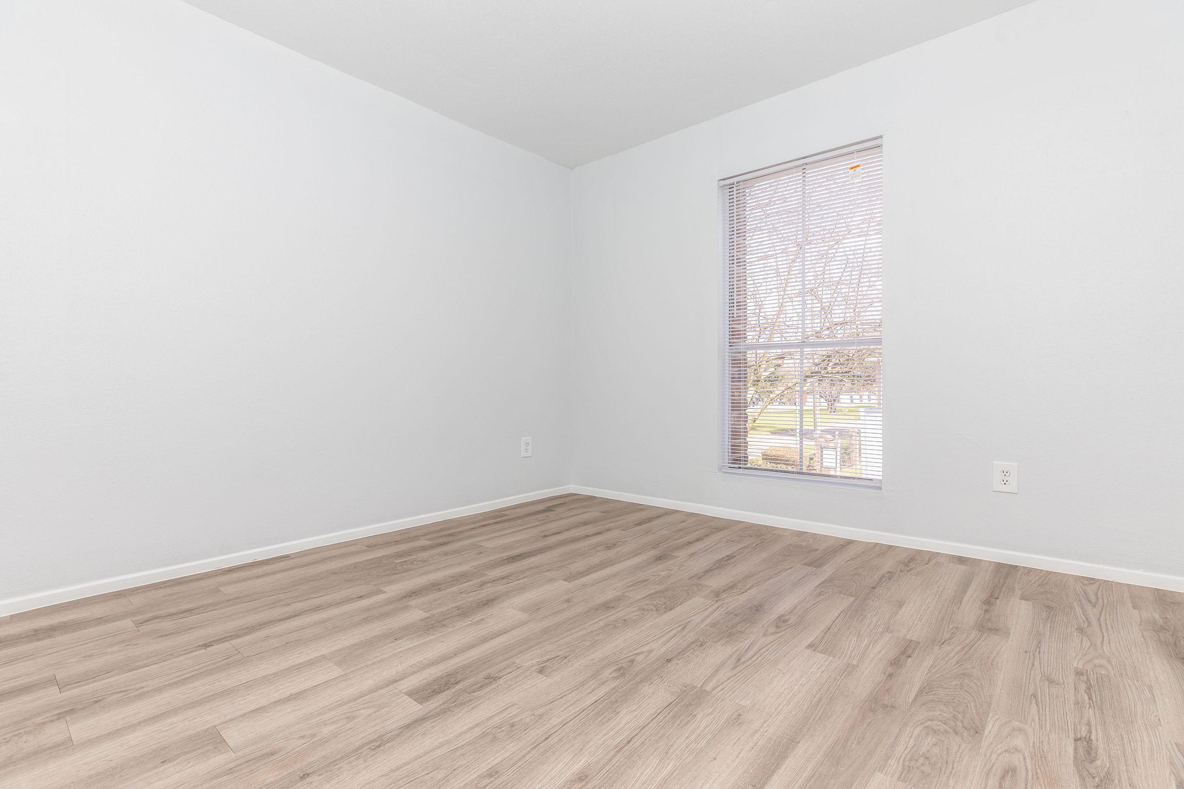 Empty room with light gray walls and light wooden flooring. A small window with blinds allows natural light to enter. The space appears clean and modern, ideal for various uses such as a living area or office.