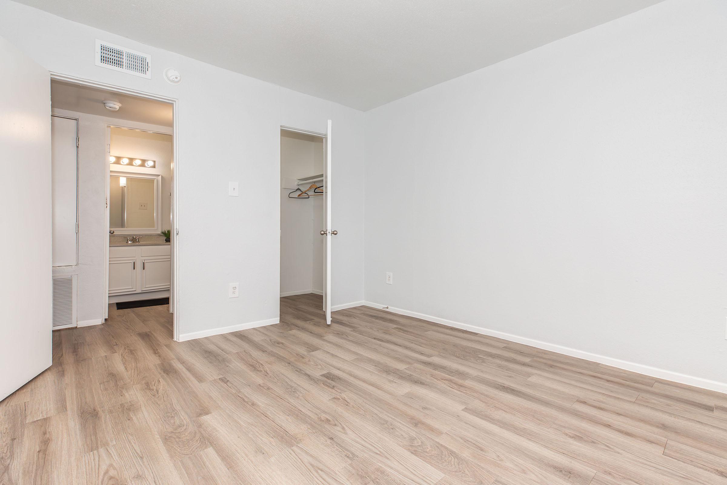 Empty room with light gray walls and laminate flooring. A door on the left leads to a bathroom area with vanity lighting visible. An open closet is on the right, showcasing hanging space. The overall atmosphere is clean and minimalistic.