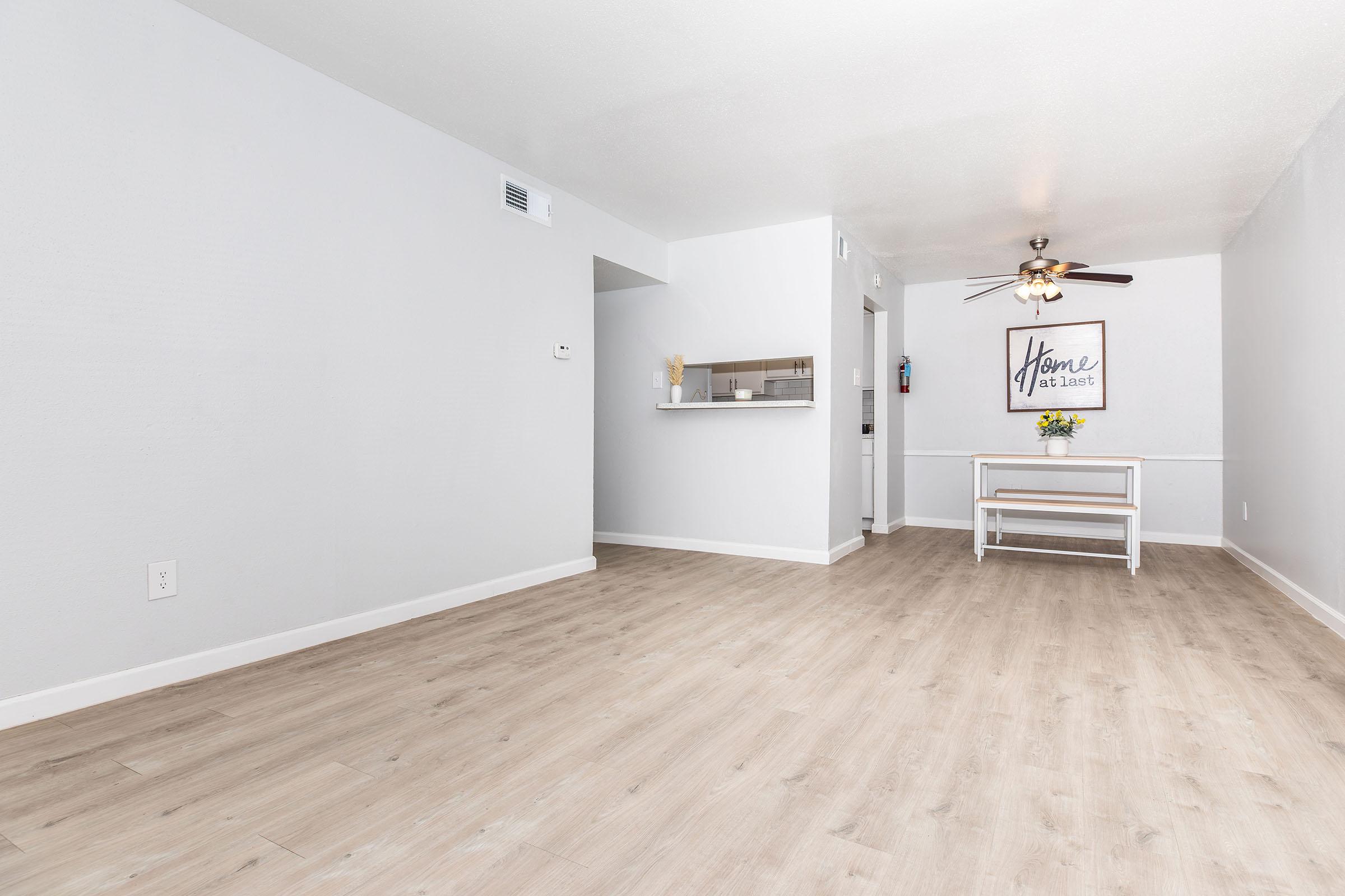 Empty living room with light wood-style flooring, pale gray walls, and a ceiling fan. A small dining area is visible in the background with a table and decorative flowers. There’s a wall sign that reads "Home." The space is bright and uncluttered, creating a welcoming atmosphere.