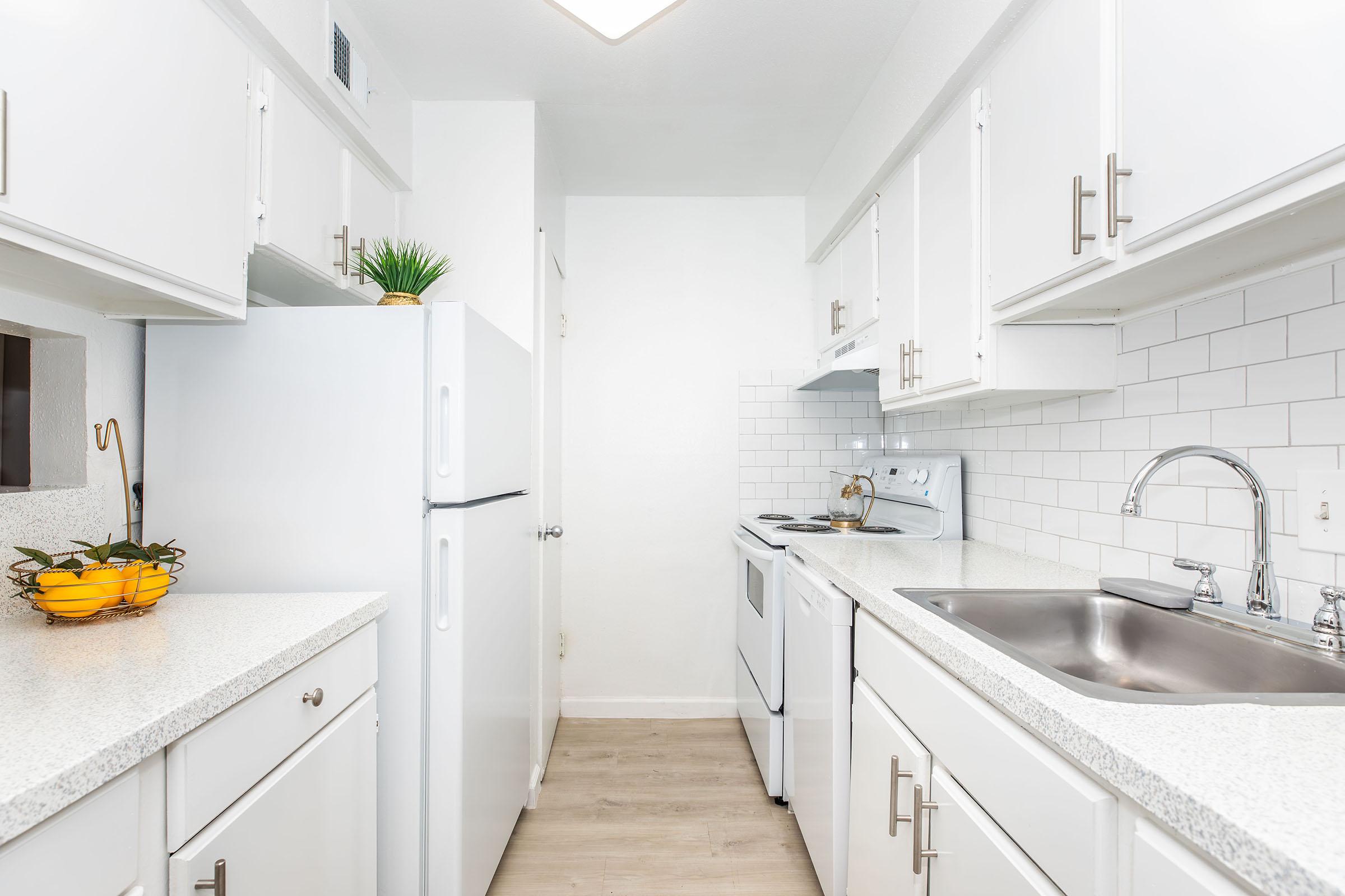 Modern kitchen featuring white cabinetry, a refrigerator, an electric stove, and a sink. The countertops are light-colored with a speckled pattern. White subway tiles adorn the backsplash, while a small plant and a bowl of oranges add a touch of greenery and color. The space is bright and well-organized.