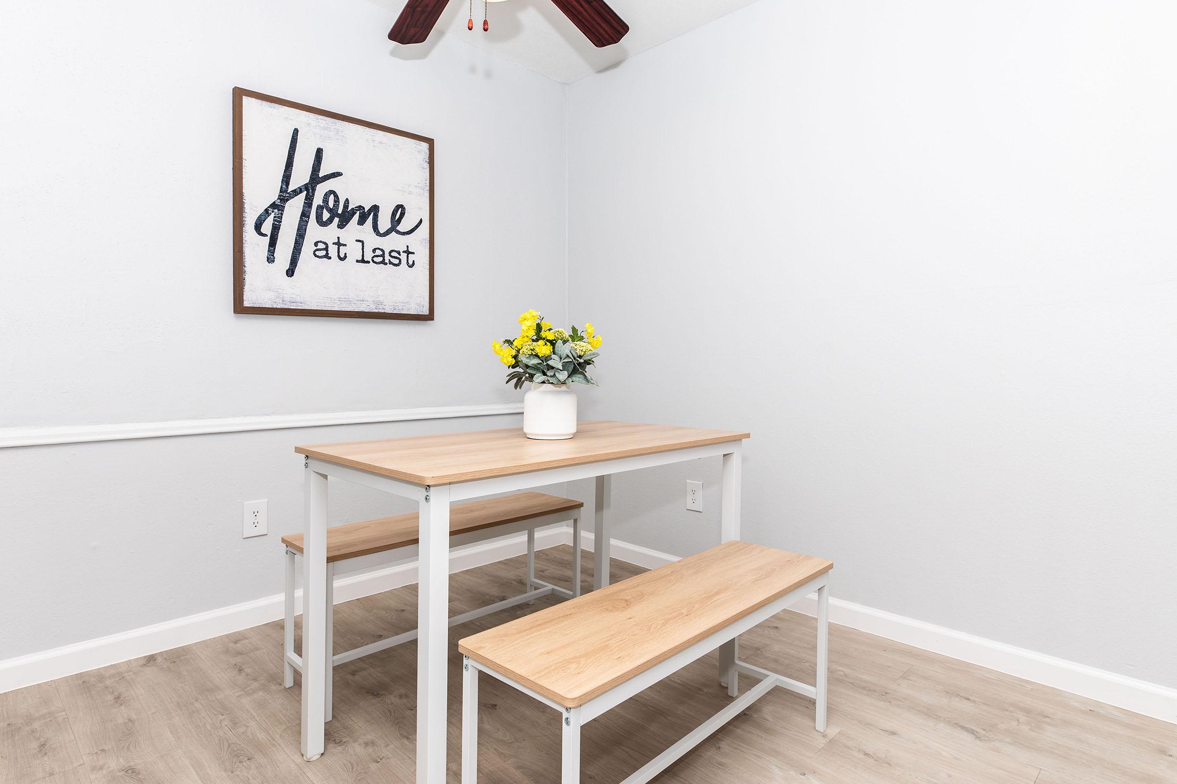 A simple dining area featuring a light wooden table and two benches, accompanied by a vase of yellow flowers. Above the table hangs a sign that says "Home at last." The room has light gray walls and a ceiling fan, creating a cozy and inviting atmosphere.