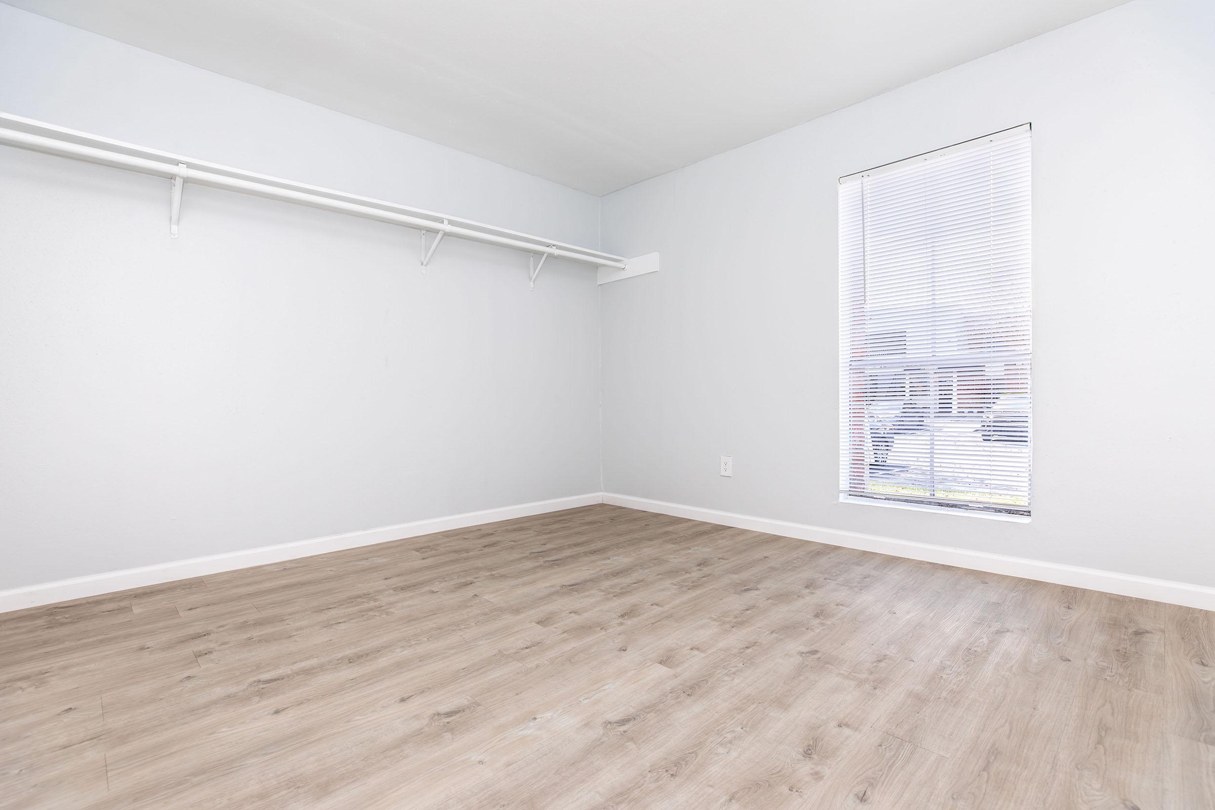 Empty room with light gray walls and a window covered by blinds. The floor is light wood laminate, and there's a white wall-mounted shelf along one side. Natural light is coming through the window, highlighting the spaciousness of the room.