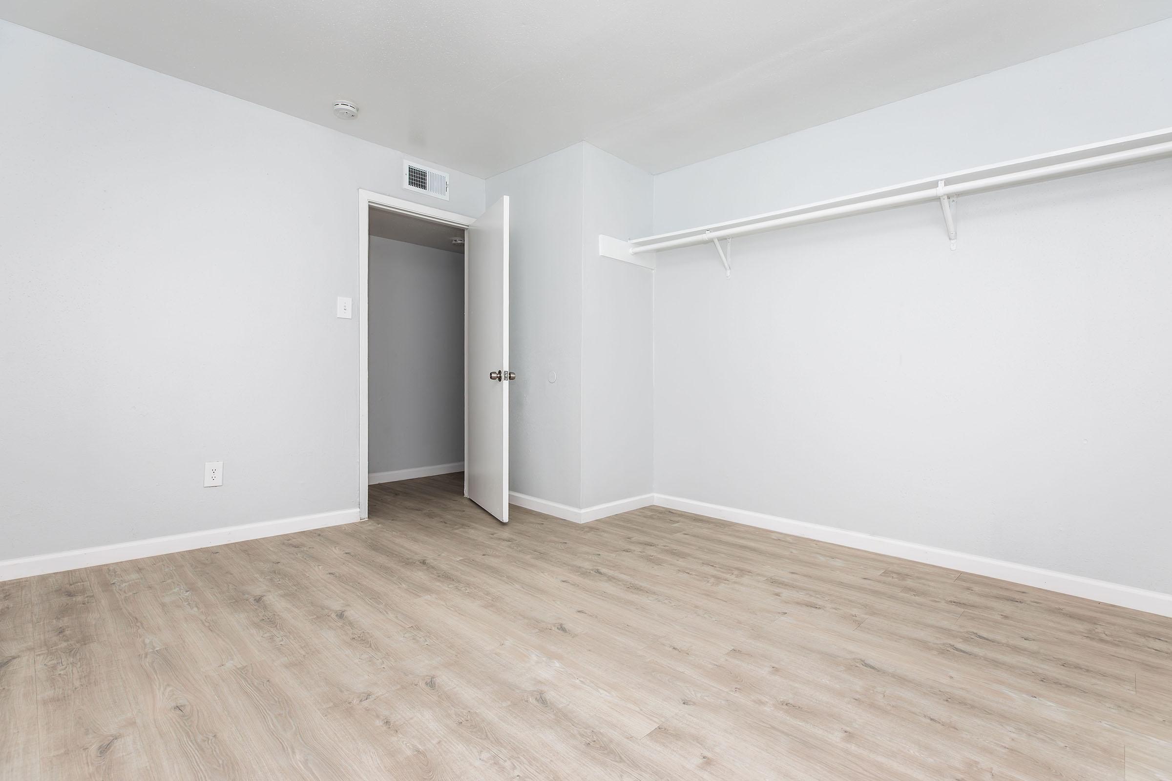 A vacant room with light gray walls and a wooden floor. There is an open white door leading to another space, and a closet rod runs along one wall. The room has a clean, minimalistic appearance, with ample natural light illuminating the area.