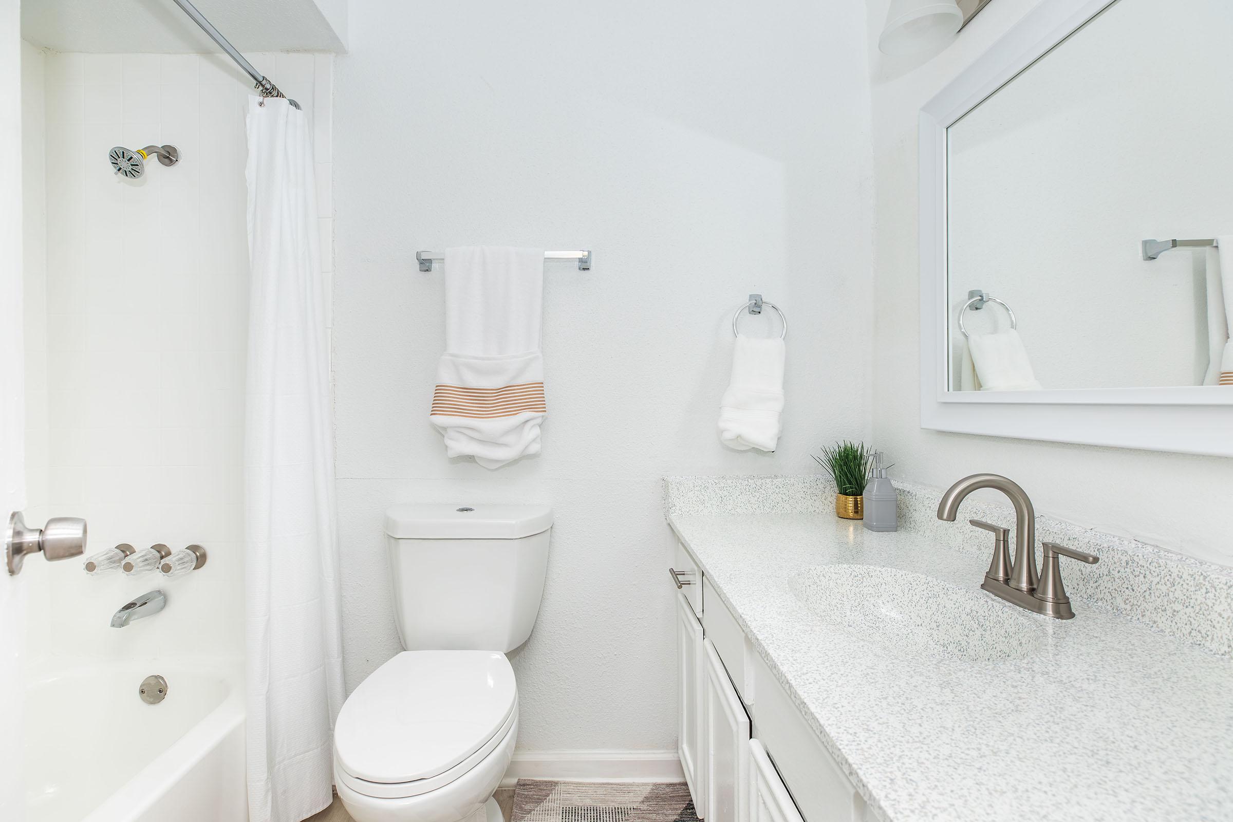 A clean and modern bathroom featuring a white bathtub with a shower curtain, a compact toilet, a sink with a granite countertop, and a large mirror. Two white towels are neatly hung on a rack, and a small potted plant adds a touch of greenery to the space. Bright walls enhance the overall light and airy feel.