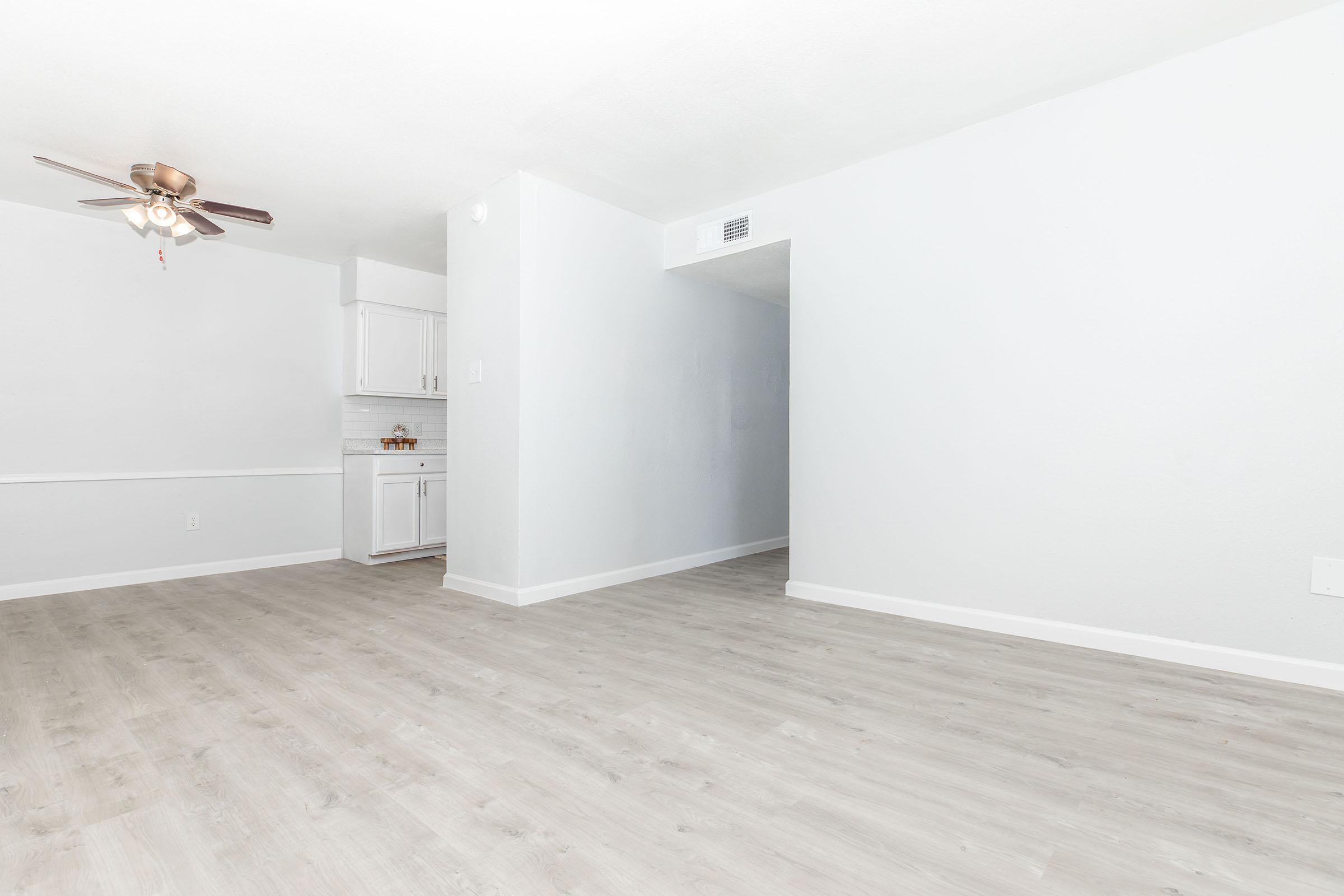 A bright and spacious room with light gray walls and wooden flooring. A ceiling fan is visible, and there is a small kitchen area with white cabinets in the background. The room is well-lit, suggesting a welcoming and airy atmosphere.
