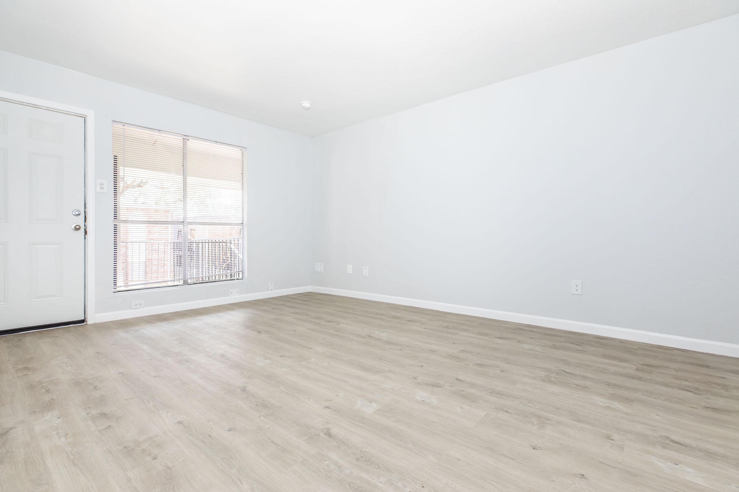 A spacious, empty living room with light-colored walls and a wooden floor. There is a large window with blinds allowing natural light to enter. A door is visible on the left side, leading outside. The room feels bright and airy, with minimal furnishings or decor.