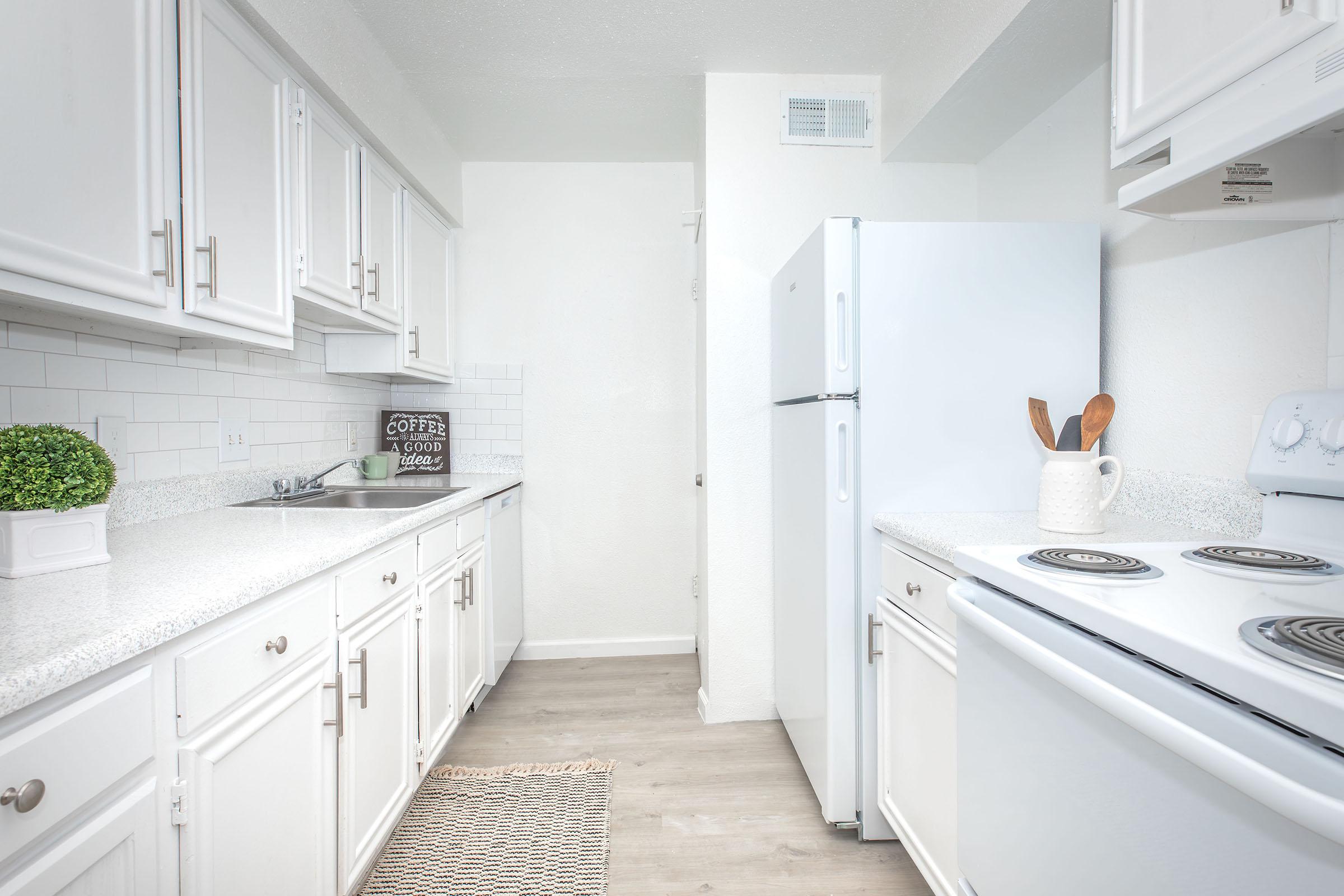 Bright and modern kitchen featuring white cabinets, a spacious countertop, and stainless steel appliances. A small decorative plant and a coffee sign add a cozy touch. The floor is light-colored laminate, and the space is well-lit with natural light, creating an inviting atmosphere.