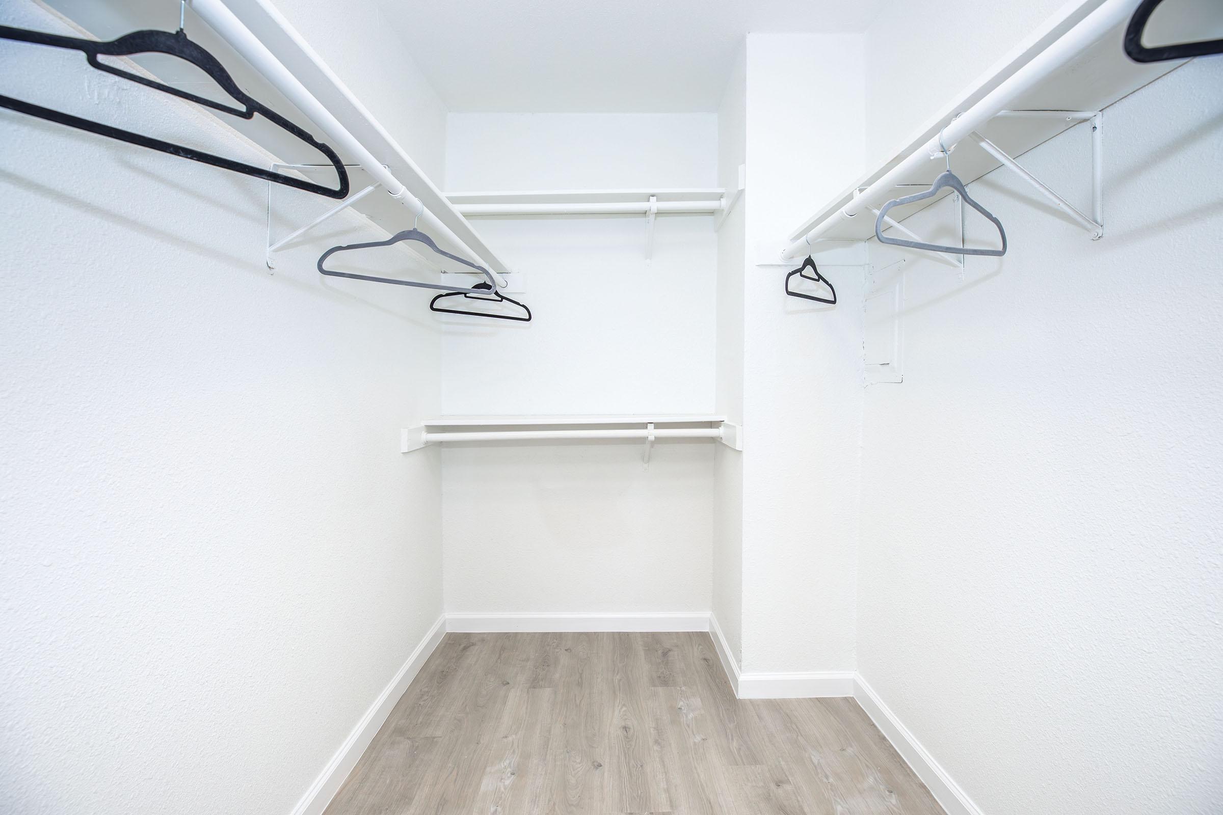 Empty closet with two hanging rods and a few black hangers. The walls are white and the floor is light-colored laminate. The space is well-lit and minimalistic, creating a clean and organized appearance.