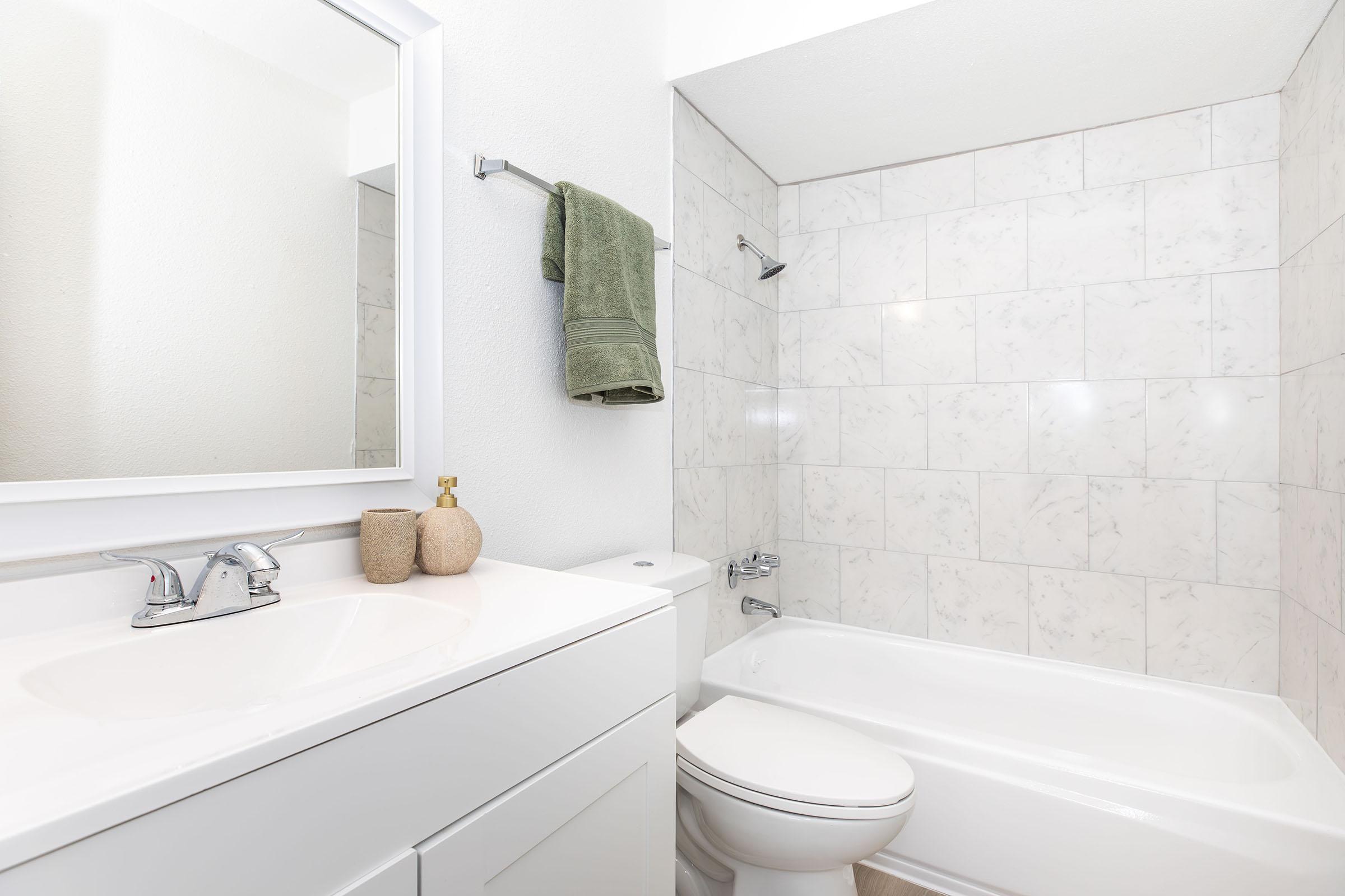 Bright and clean bathroom featuring a white bathtub, modern sink with a sleek faucet, and a large mirror. The walls are tiled with light-colored marble, and a green towel hangs on a rack. A small decorative item and a soap dispenser are placed on the counter.