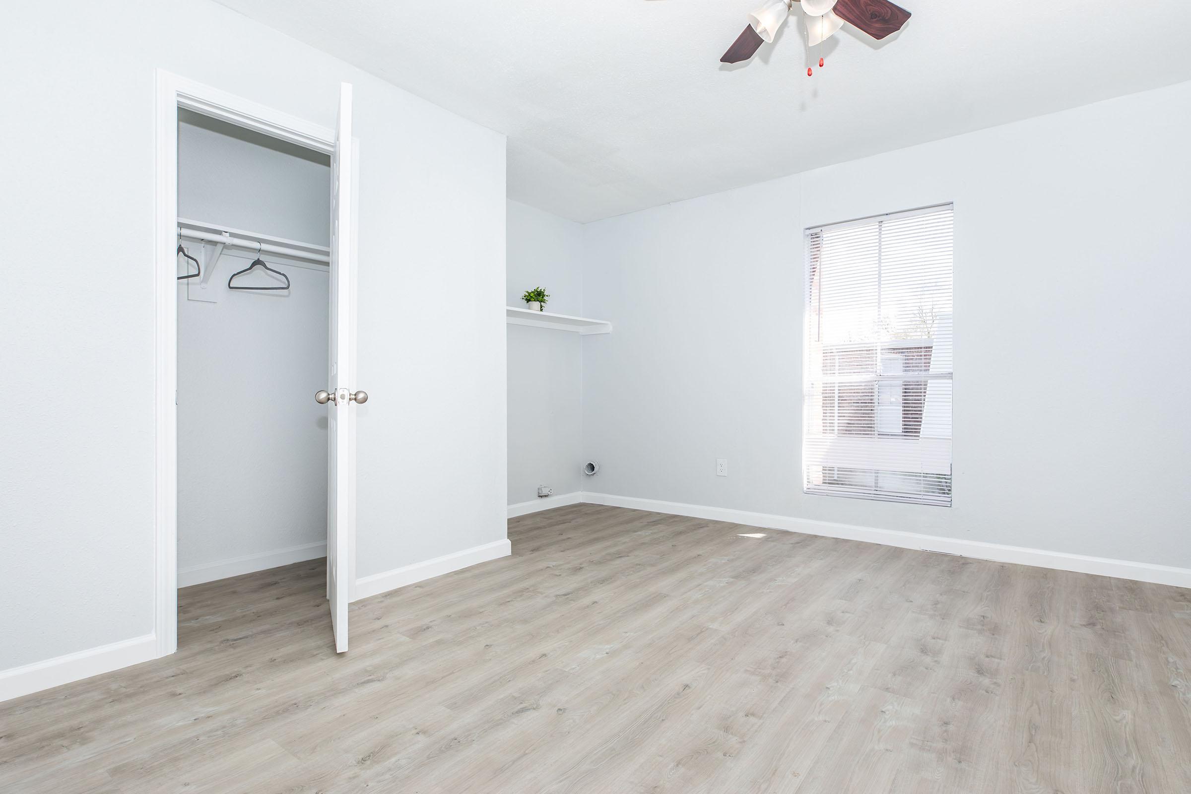 Empty room with light gray walls and a wooden floor. A white door is slightly open, revealing a closet with hangers. There's a small shelf on the wall and a window with blinds that lets in natural light. A ceiling fan is visible above.