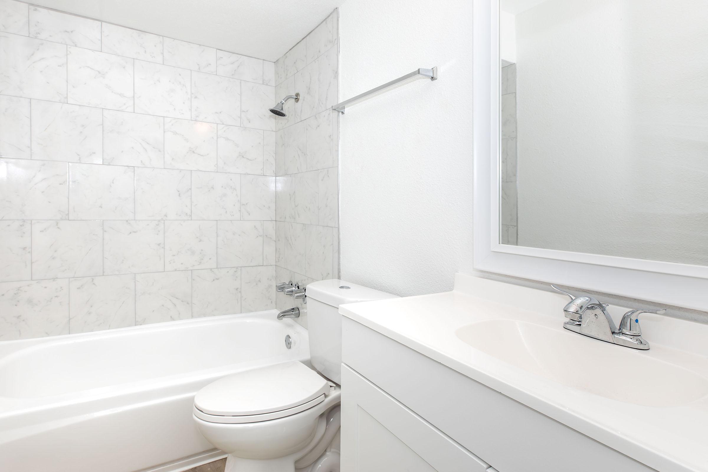 A modern bathroom featuring a white tub, toilet, and sink with a minimalist design. The walls are adorned with light-colored marble tile, and there is a large mirror above the sink. Natural light illuminates the space, creating a clean and airy atmosphere.