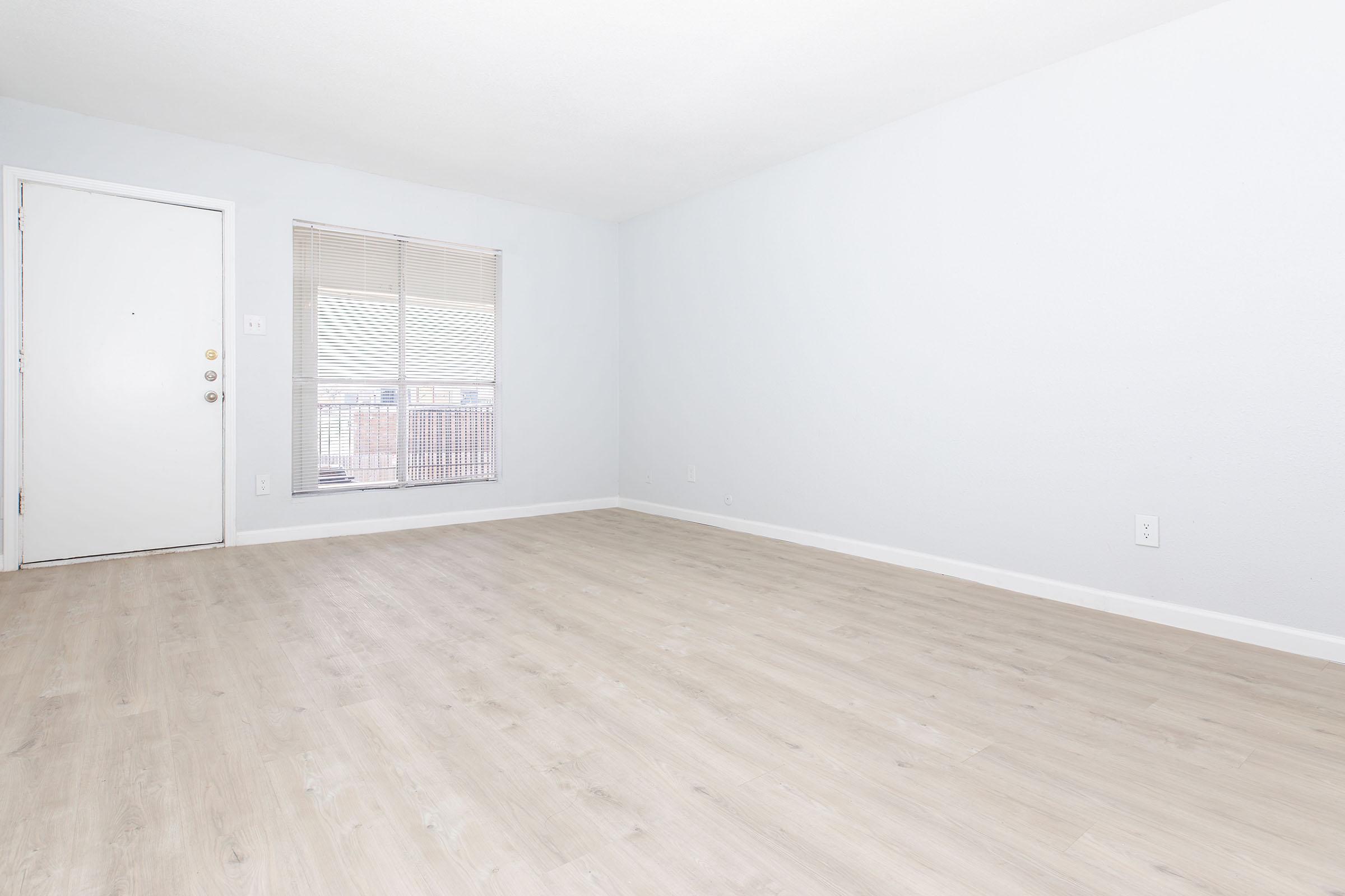 A bright and empty room with light-colored walls and a light wood floor. There is a closed door on the left and a window with blinds on the right, allowing natural light to enter. The space appears clean and ready for furnishings.