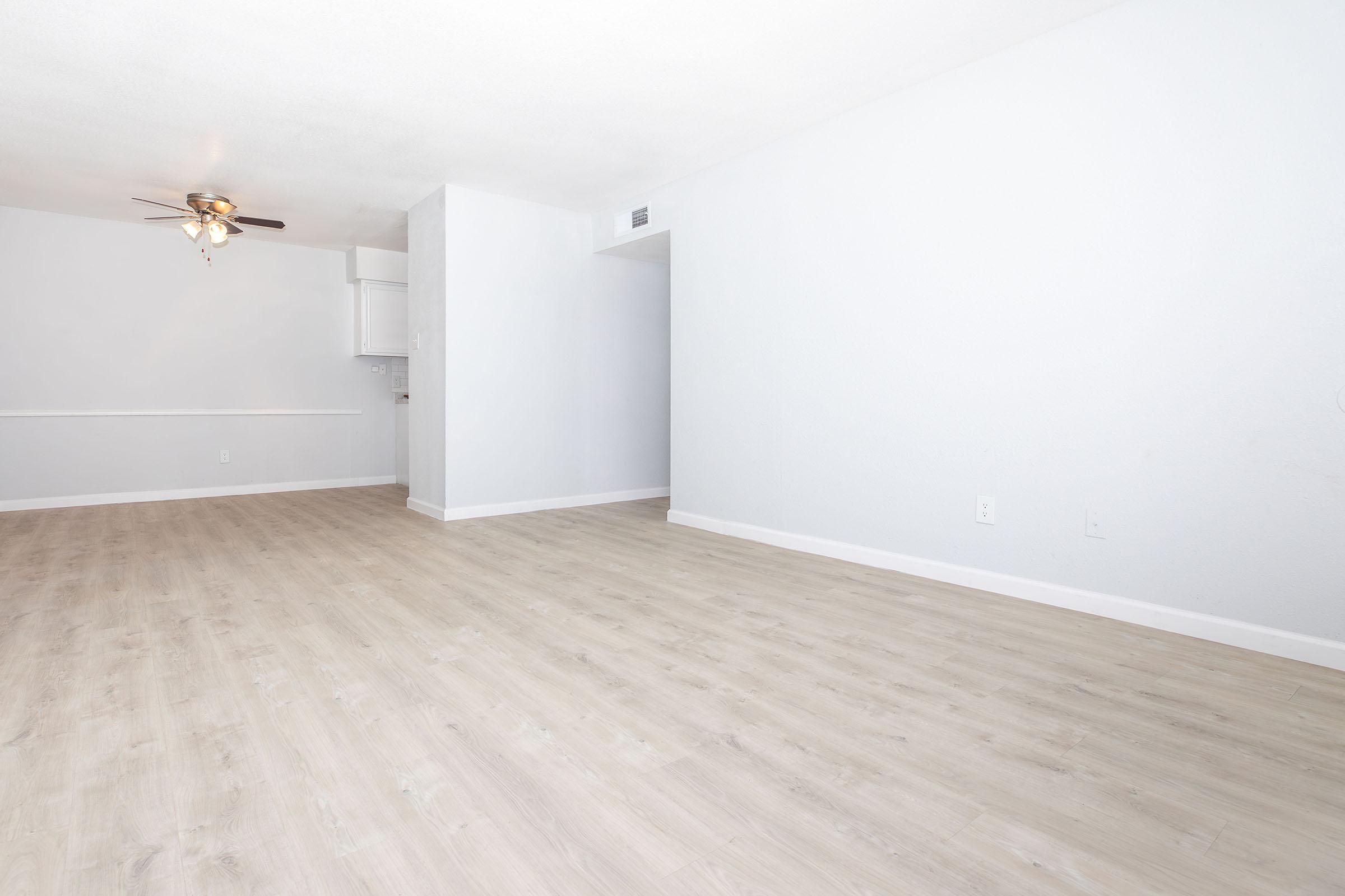 Bright and airy interior of a vacant apartment, featuring light-colored walls and light wood-style flooring. A ceiling fan is visible in the corner, and there is an open space leading to a kitchenette on the left. The room appears spacious and inviting, making it suitable for various furnishings and decorations.