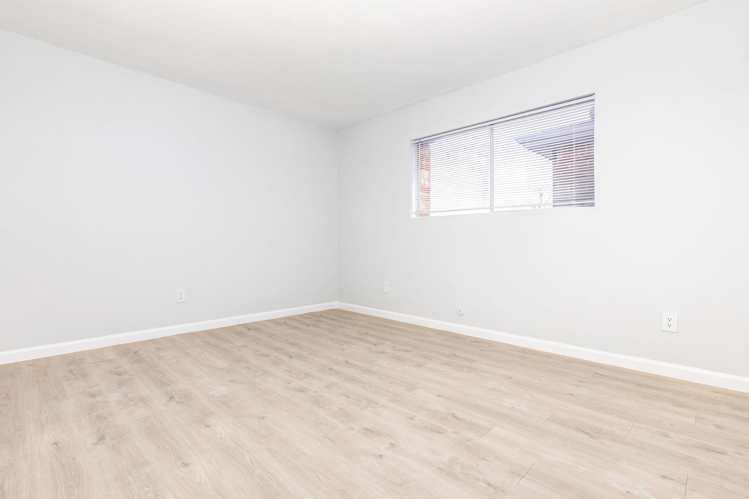 A blank, empty room with light gray walls and a light wood floor. A small window with blinds allows natural light to enter. The space is devoid of furniture or decorations, creating a minimalist appearance.