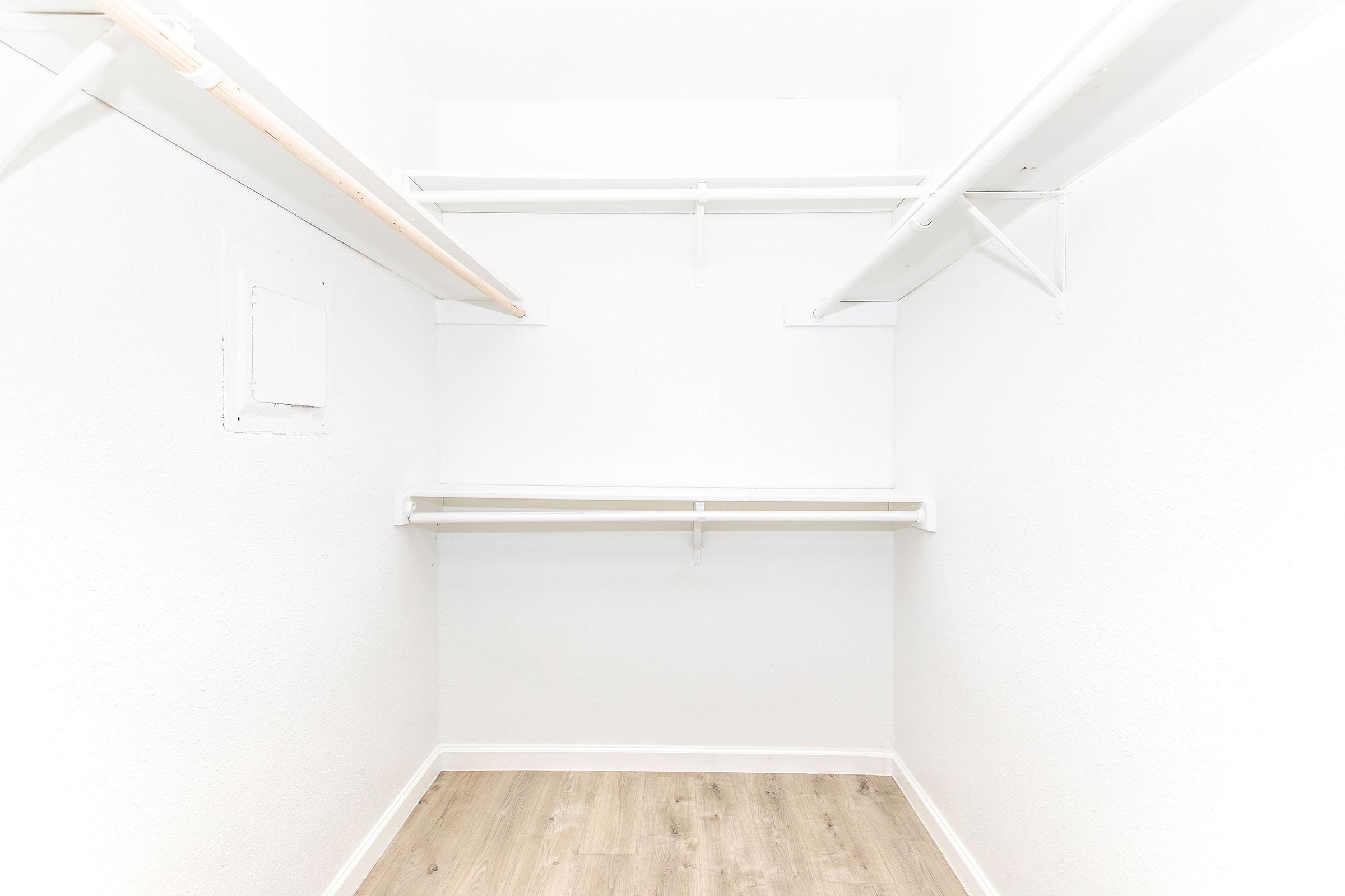 A spacious, empty closet with white walls, a wooden floor, and two hanging rods. There are shelves along the sides, providing ample storage space. The overall aesthetic is bright and clean, with natural light enhancing the uncluttered appearance.