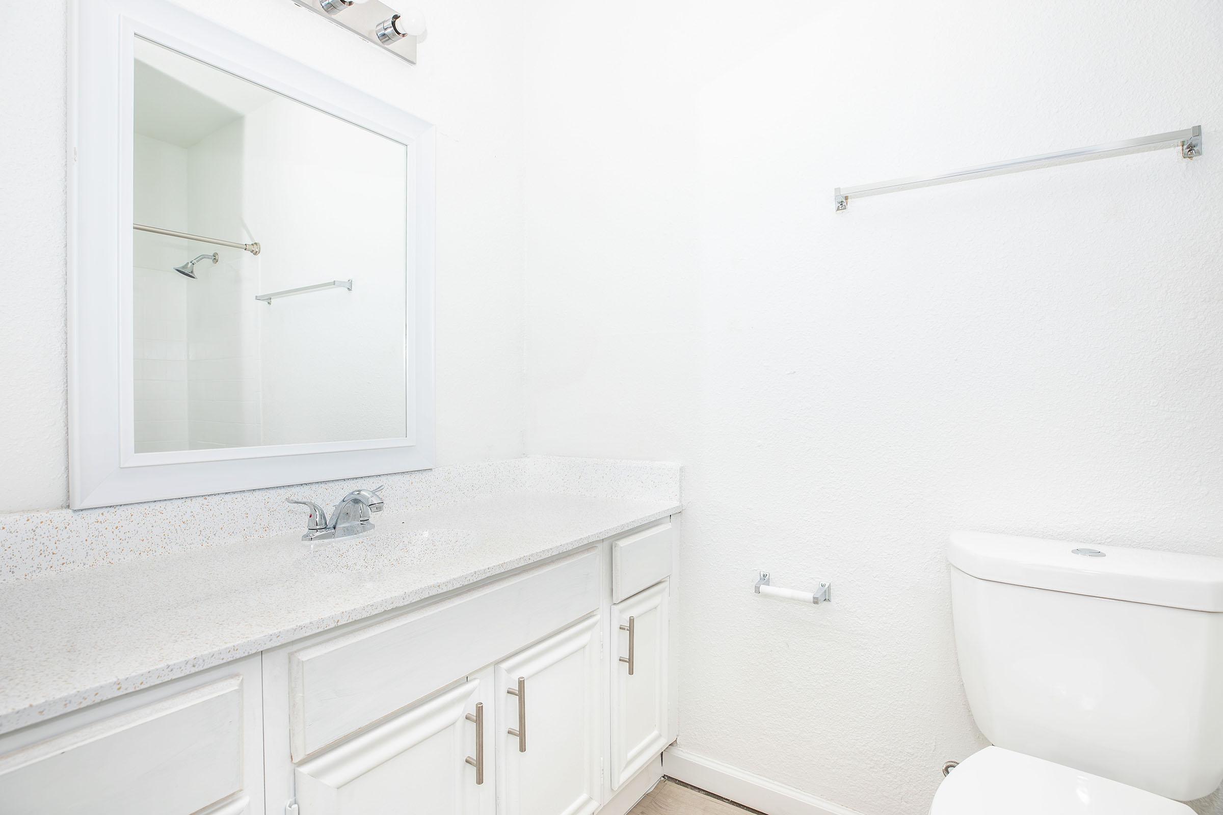 Bright and minimalistic bathroom featuring a white vanity with a granite countertop, a rectangular mirror above it, and a clean white toilet. The walls are painted white, and there's a towel bar mounted on the wall, creating an open and airy atmosphere.
