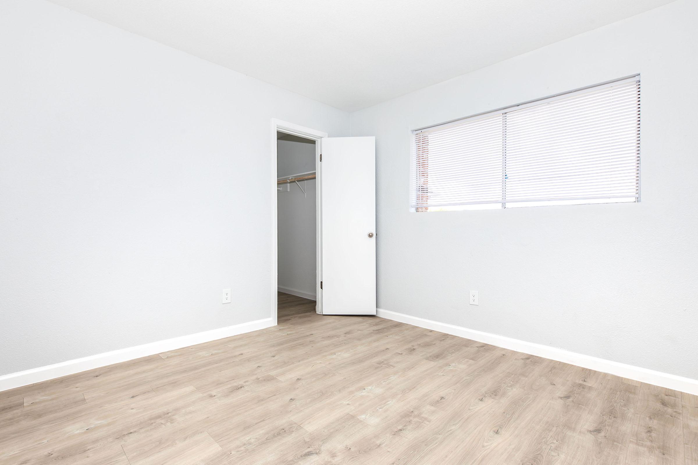 A minimalist bedroom featuring light gray walls and wooden flooring. There is a window with blinds allowing natural light, and a closet with an open door on the left side. The room is unfurnished, giving it a spacious and airy feel.