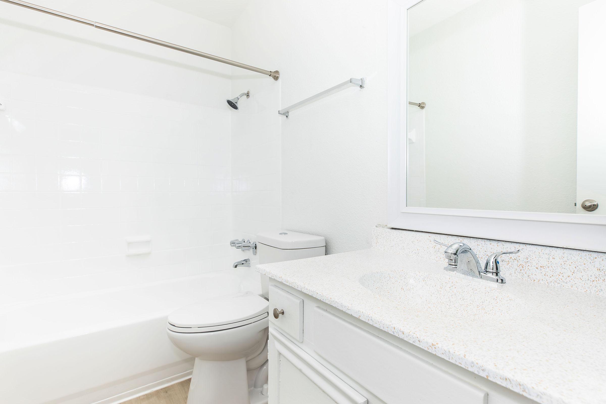 Bright, clean bathroom featuring a white tiled shower and tub, a modern toilet, and a stone countertop with a sink. A rectangular mirror is mounted above the sink, and there are minimalistic fixtures and a towel bar on the wall. The overall design is simple and contemporary, with light-colored walls.