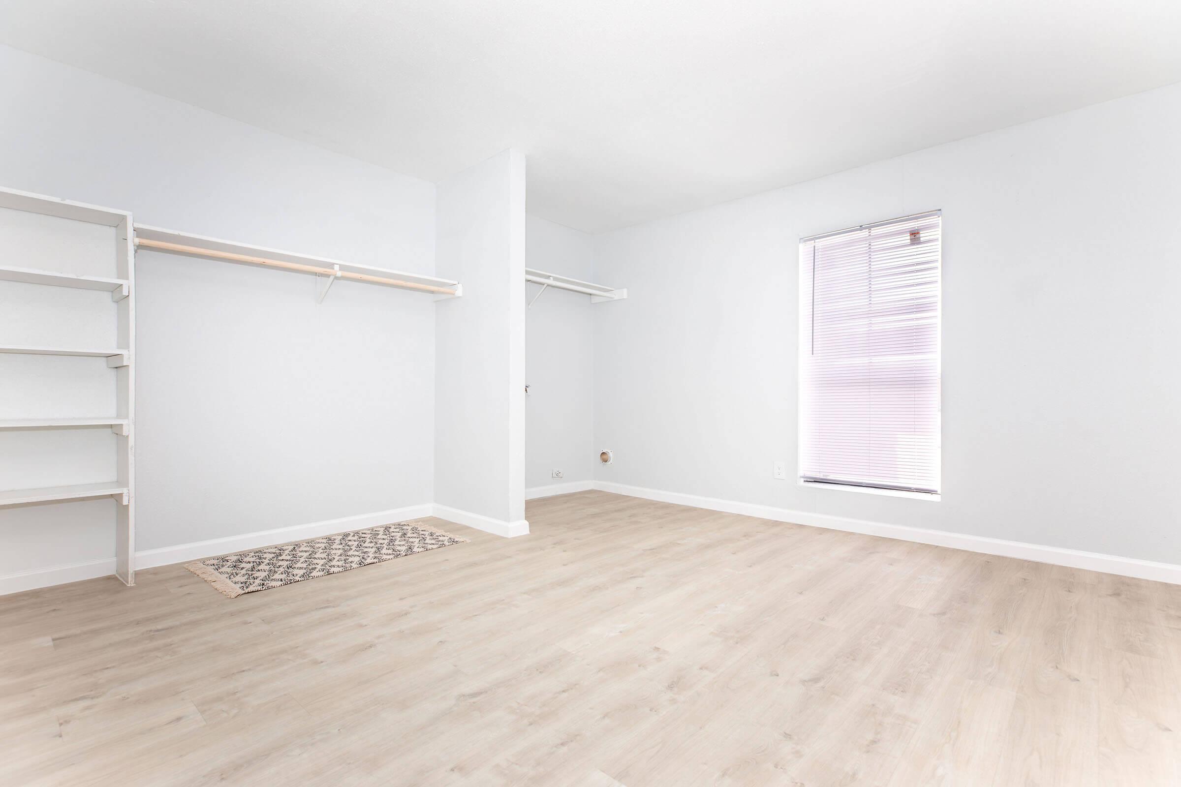 A spacious, empty room with light-colored walls and wooden flooring. The room features a window with blinds, a small area rug on the floor, and a shelf on one wall with a hanging rod for clothing. The overall aesthetic is bright and minimalist, ideal for various decor styles.
