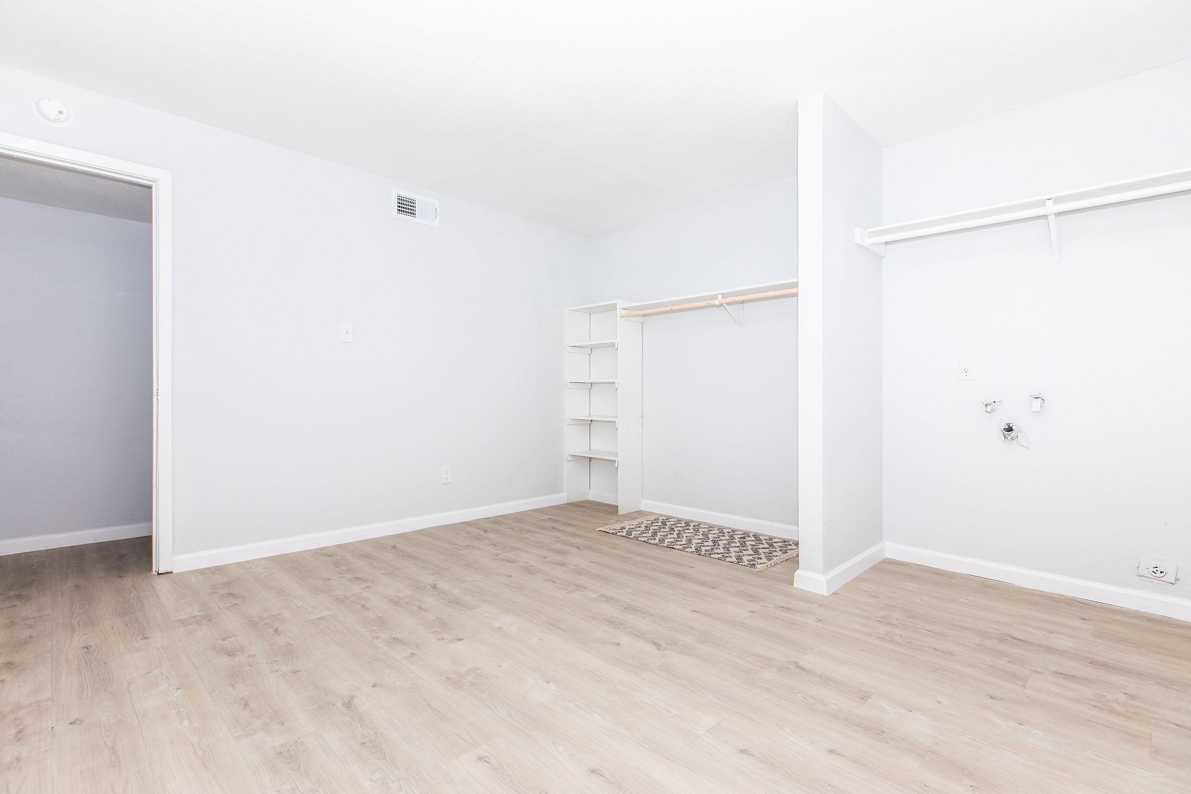 A spacious, empty room with light gray walls and a light wooden floor. It features a small closet nook with a hanging rod, a shelf unit against one wall, and a patterned rug on the floor. There are no furnishings, creating a clean and open atmosphere.