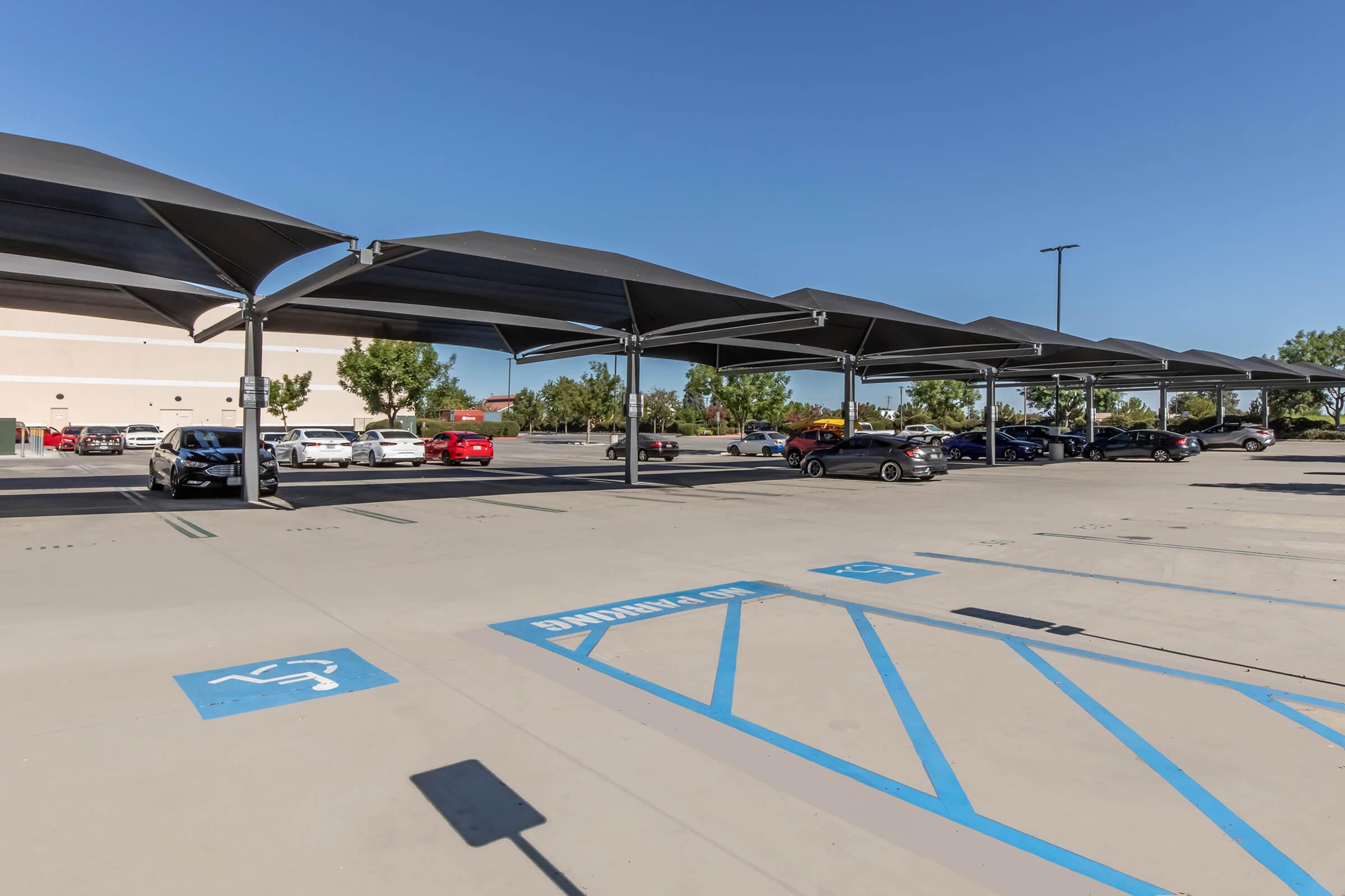 A wide view of a partially occupied parking lot featuring several covered spaces. Clear blue skies and trees in the background suggest a sunny day. Parking spaces are marked, including those designated for disabled parking. A shopping center is visible in the distance.