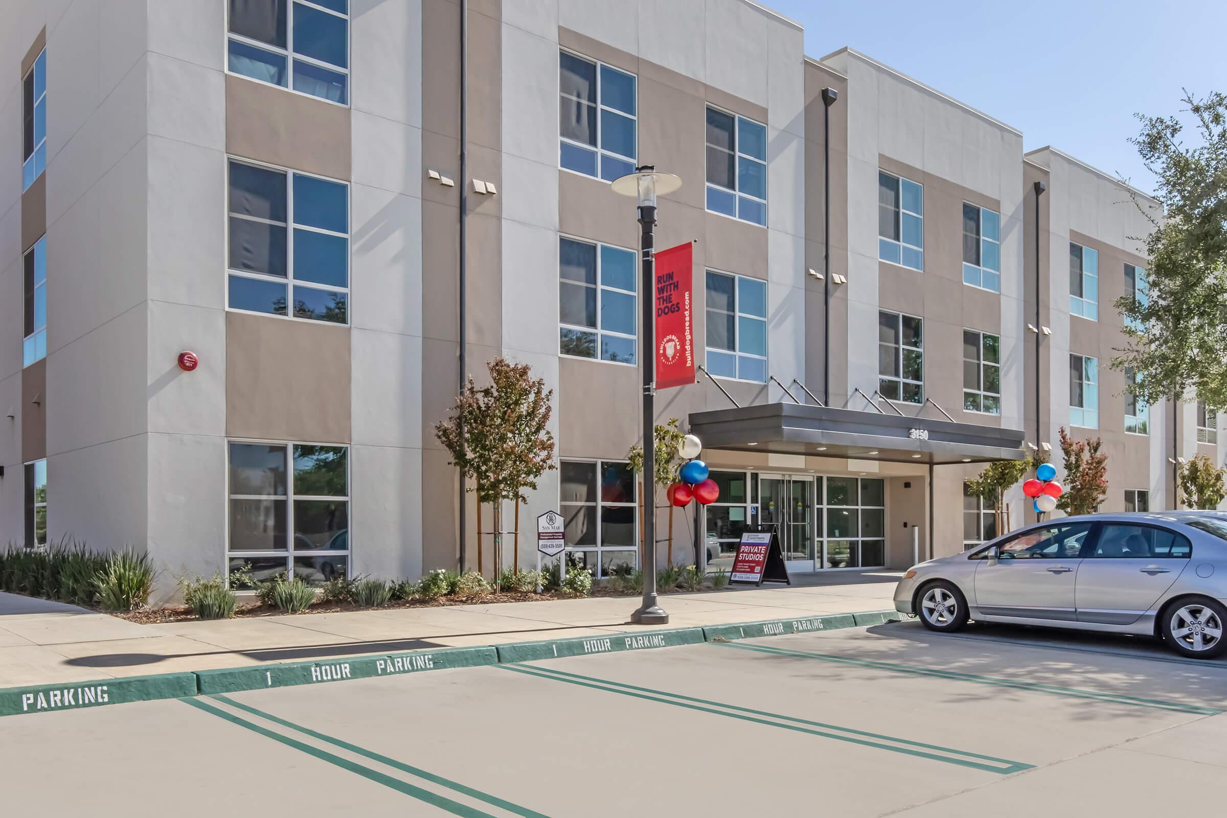 Modern building exterior with large windows and a welcoming entrance. Red, white, and blue balloons decorate the entrance. A silver car is parked in front, with designated parking lines visible. There are trees and shrubs landscaping the area. A banner hangs above the entrance.
