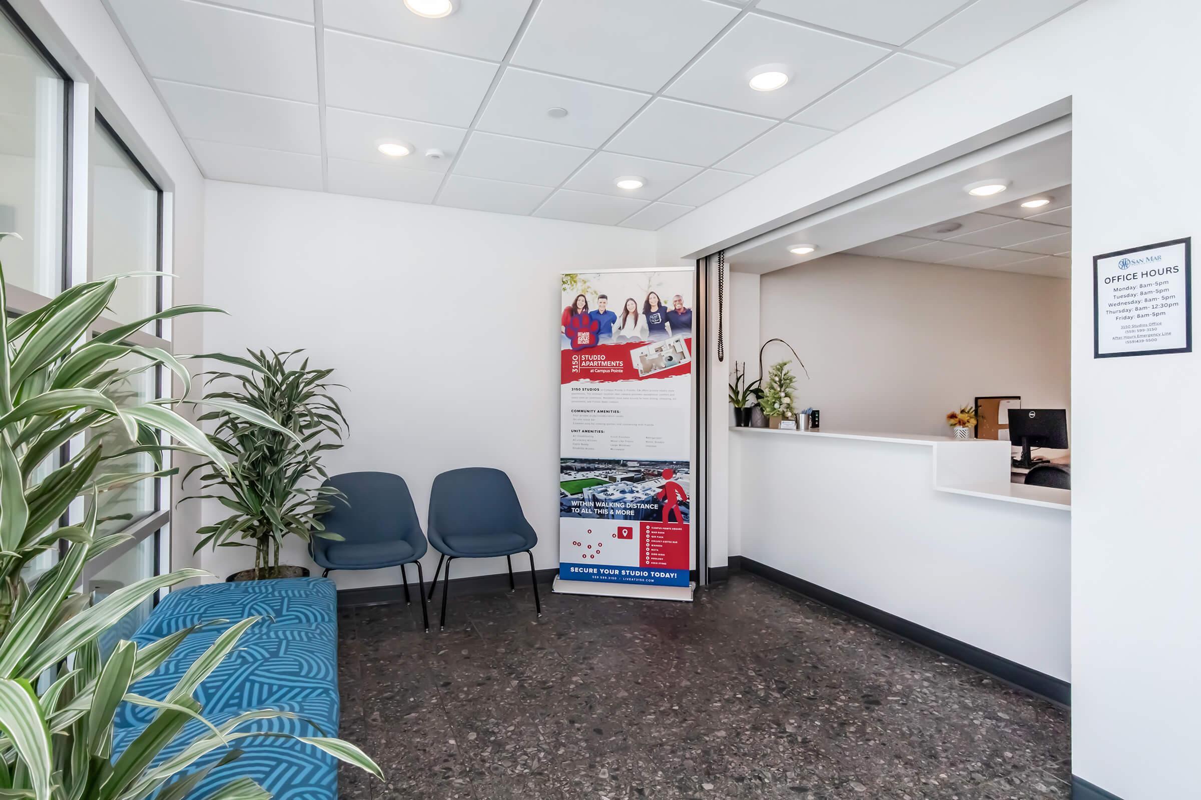 A modern office reception area featuring a blue cushioned bench, two chairs, and a reception desk. A banner displaying images of diverse people and informational content is positioned in the background. There are plants in the room, and the walls are painted in neutral colors, creating a welcoming atmosphere.