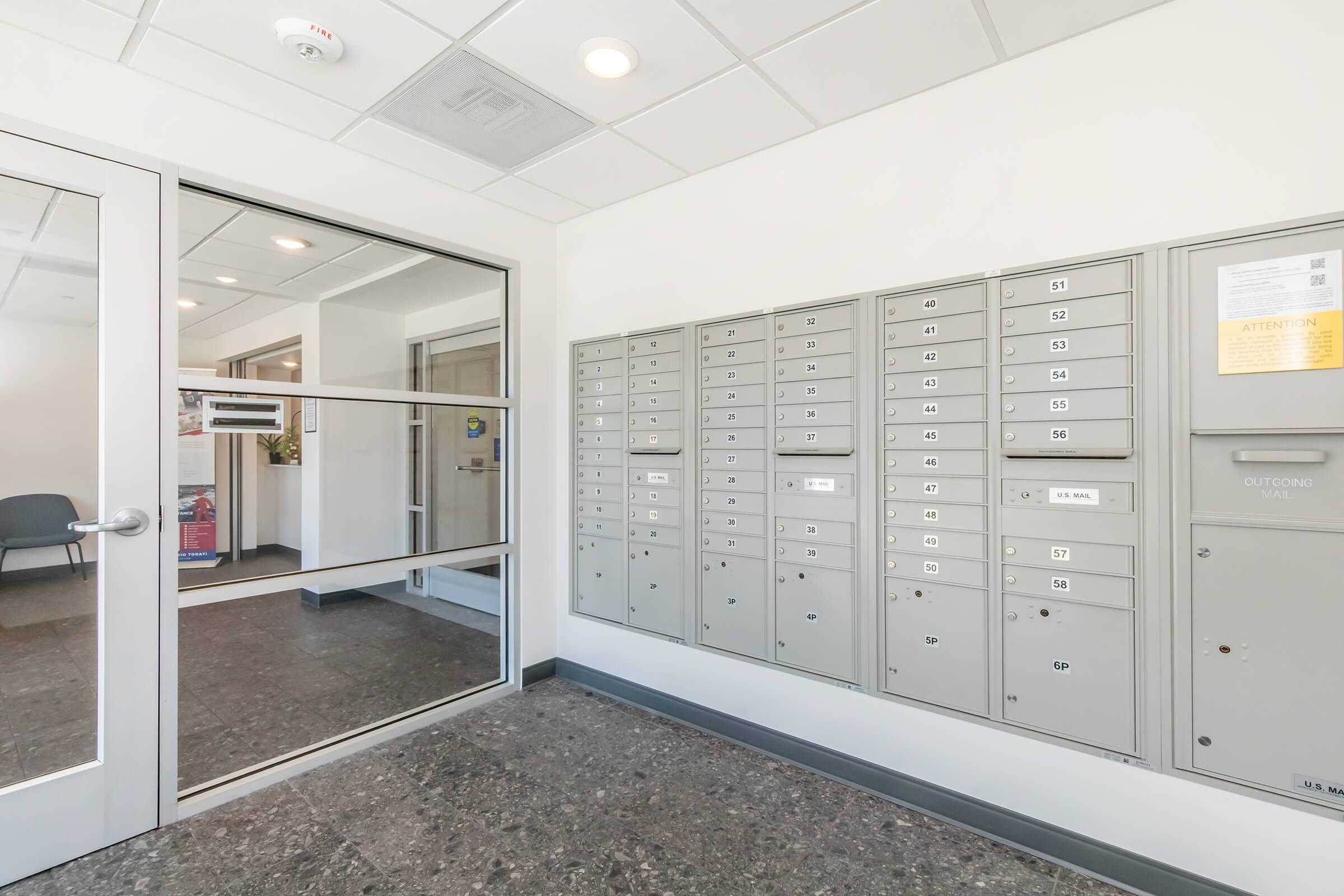 Interior view of a mailroom featuring a glass door and a row of organized metallic mailboxes. The mailboxes are mounted on a wall and numbered. The floor is made of dark tiles, and there is a chair visible in the background, suggesting a waiting area. Natural light illuminates the space.