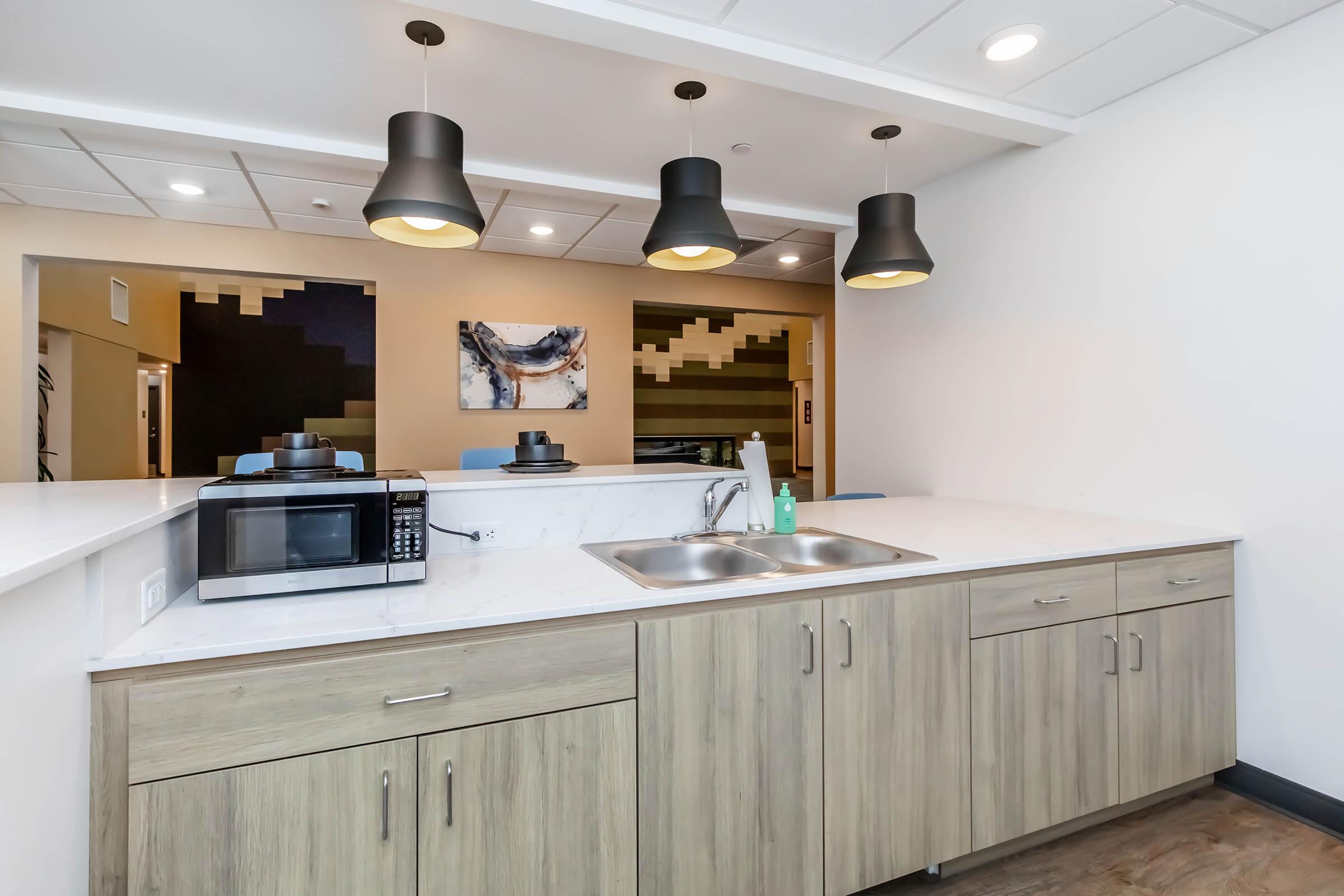 A modern kitchenette with a stainless steel sink, microwave, and wooden cabinetry. The space features sleek lighting fixtures hanging from the ceiling and a minimalist design, with a colorful abstract painting on the wall in the background. The overall aesthetic is contemporary and functional.
