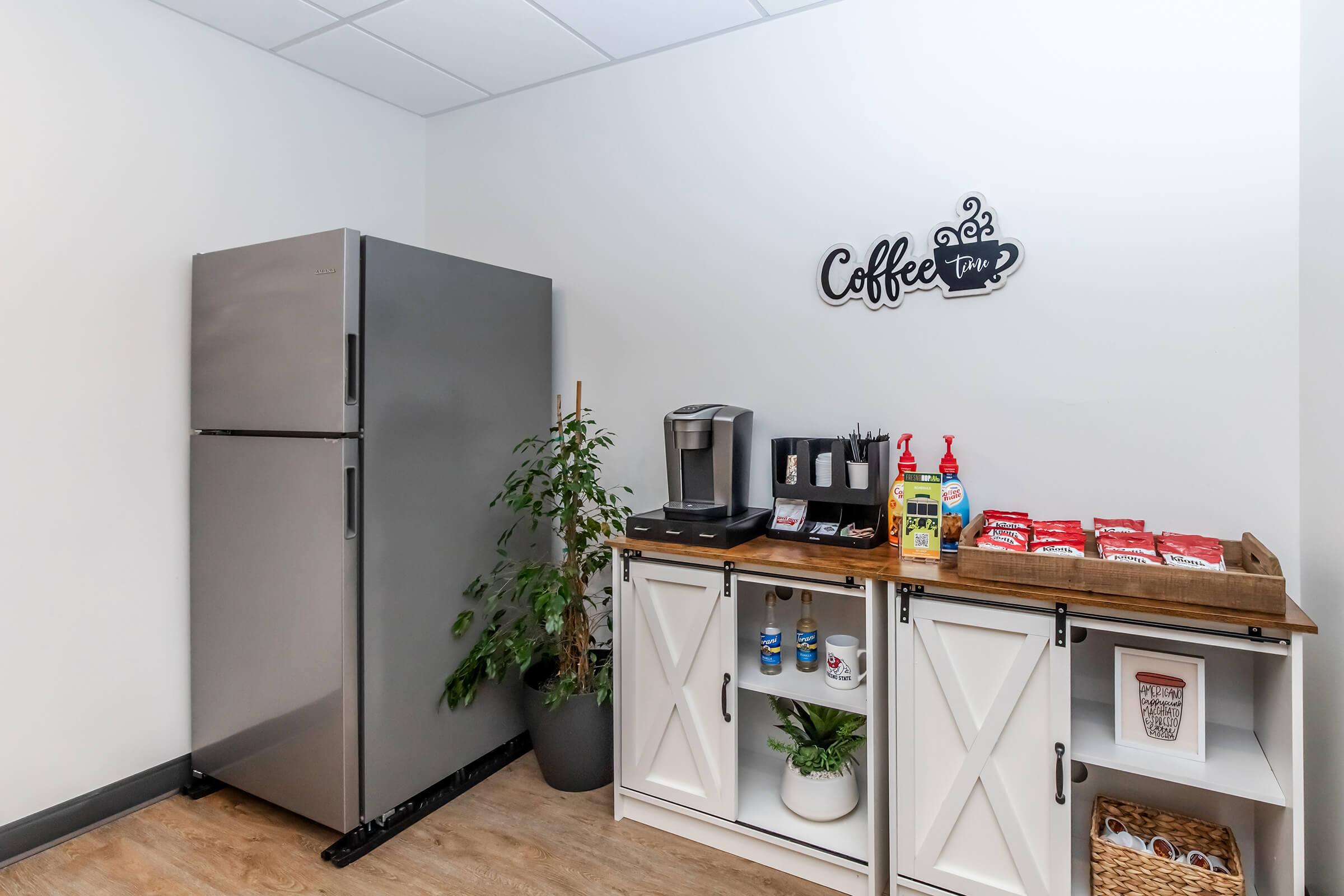 A modern kitchen area featuring a stainless steel refrigerator, a coffee maker, and a neatly organized shelf with snacks and beverages. A decorative sign that says "Coffee Time" adds a cozy touch to the space, complemented by a small potted plant. The flooring is light wood, giving a warm ambiance.