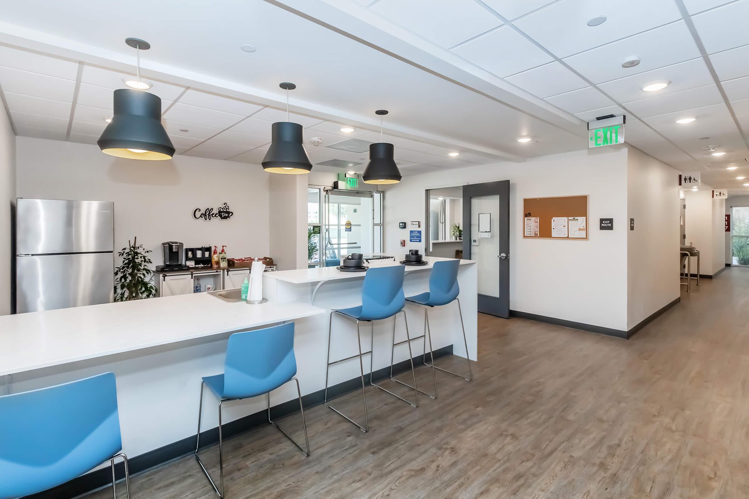 A modern office kitchen featuring a white countertop with blue chairs, pendant lighting, and a refrigerator. Coffee and supplies are visible on the counter, and a doorway leads to another area. The floor is wood-like, and the walls are bright and well-lit, creating a welcoming atmosphere.
