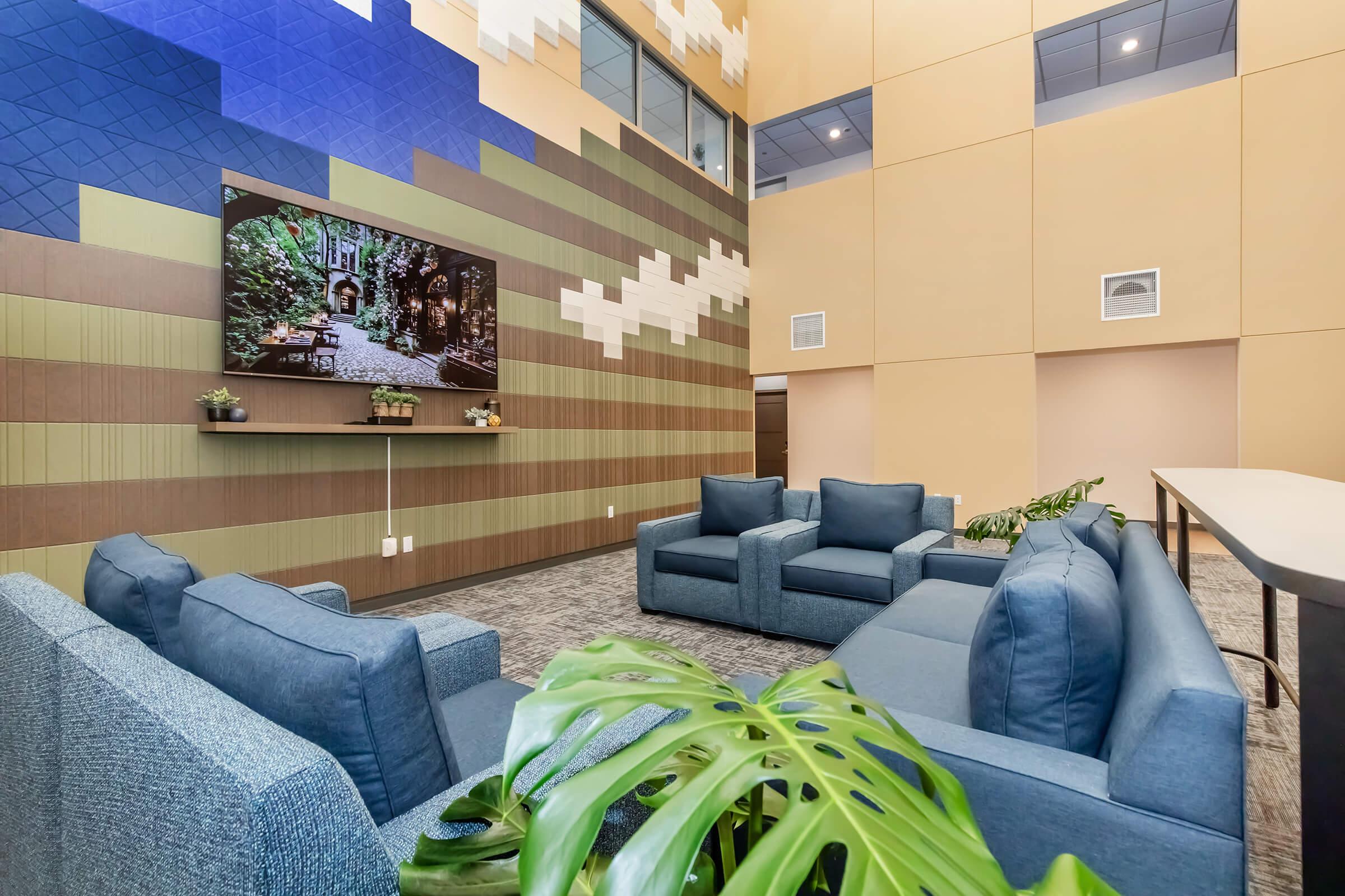 Modern lounge area featuring blue upholstered sofas arranged around a low table, with a large wall-mounted television displaying a scenic image. The decor includes green plants and textured wall designs in neutral tones, creating a welcoming and comfortable atmosphere. Natural light fills the space from large windows.