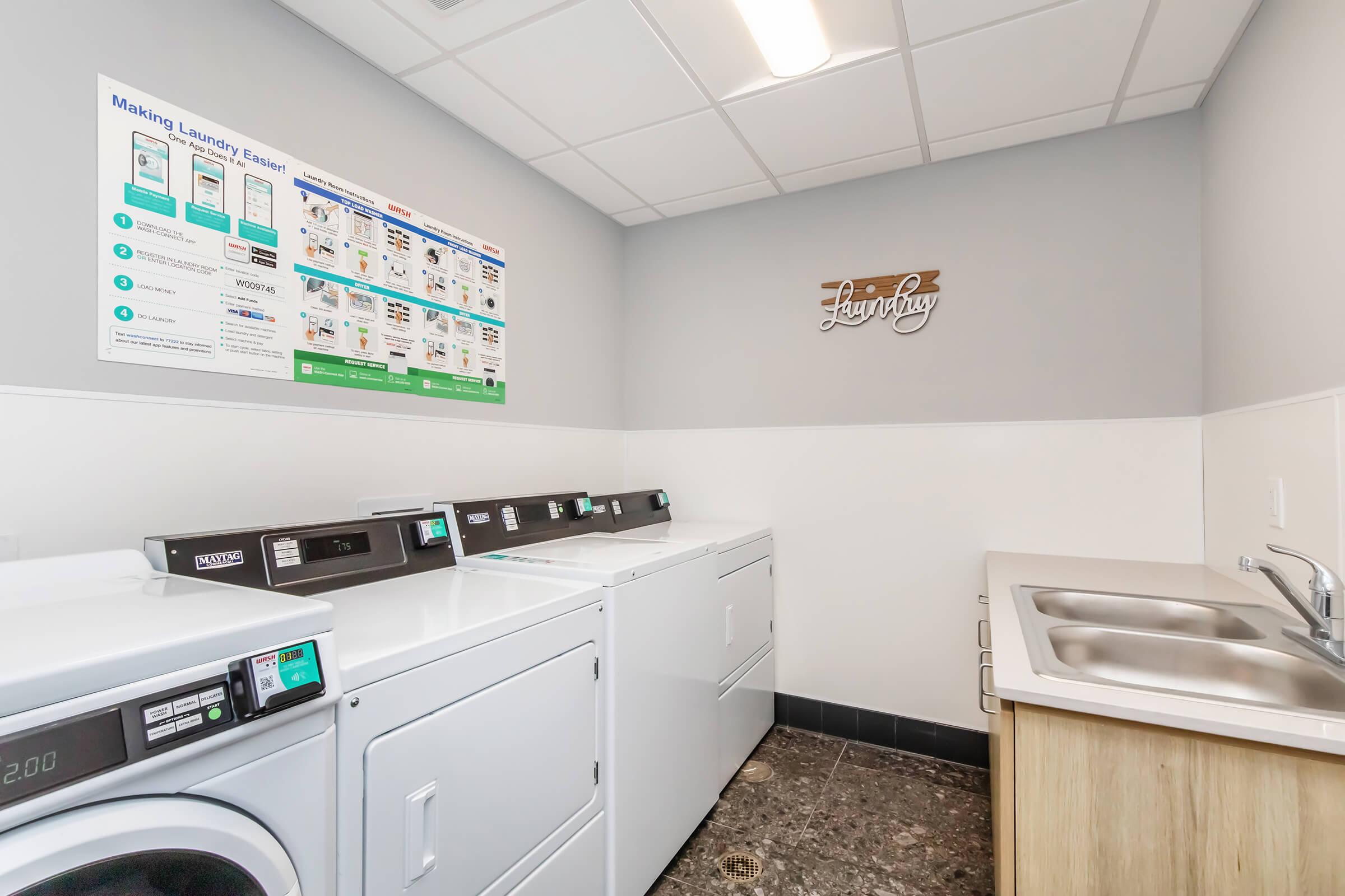 A clean and modern laundry room featuring multiple washing machines and dryers, a laundry sink, and a wall-mounted sign that says "Laundry." A bulletin board with laundry instructions and tips is displayed above the machines. The room has a neutral color scheme and good lighting.