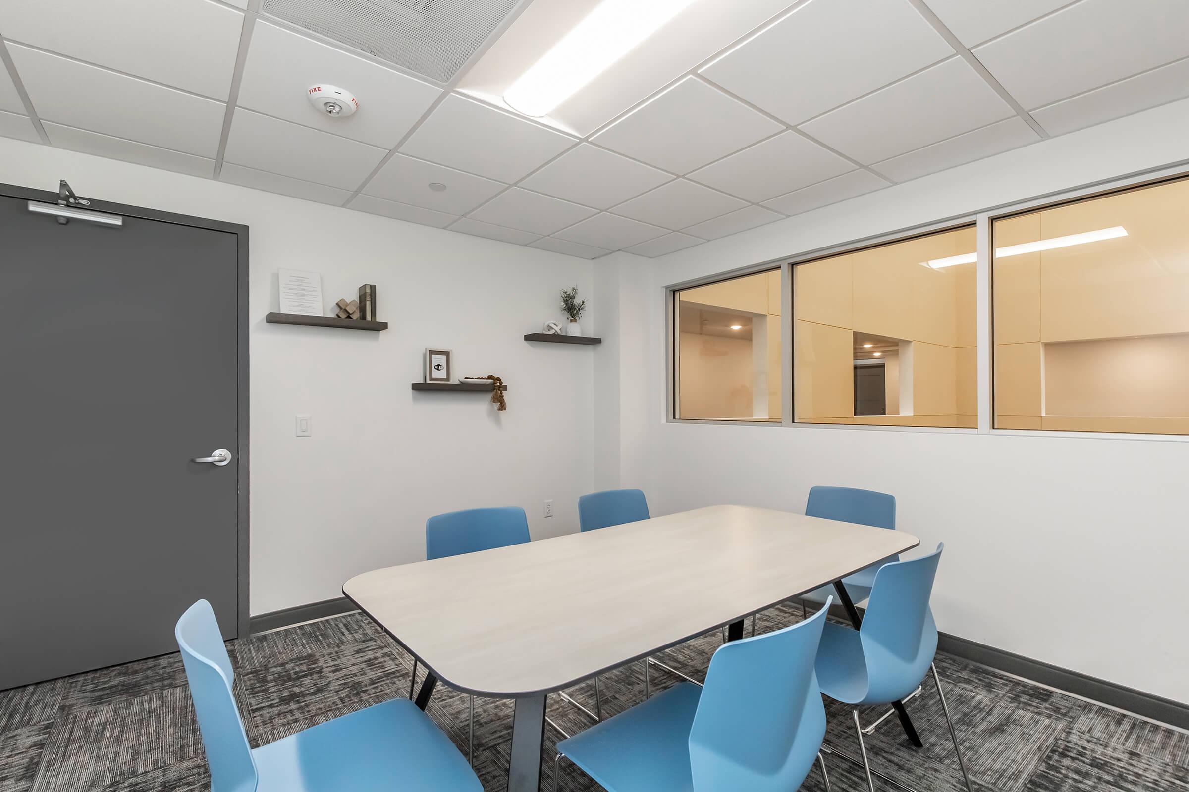 A modern conference room featuring a light-colored table with six blue chairs, a gray door, and two shelves on the wall with decorative items. Large windows provide natural light, and the walls are painted in neutral tones, creating a professional and inviting atmosphere.