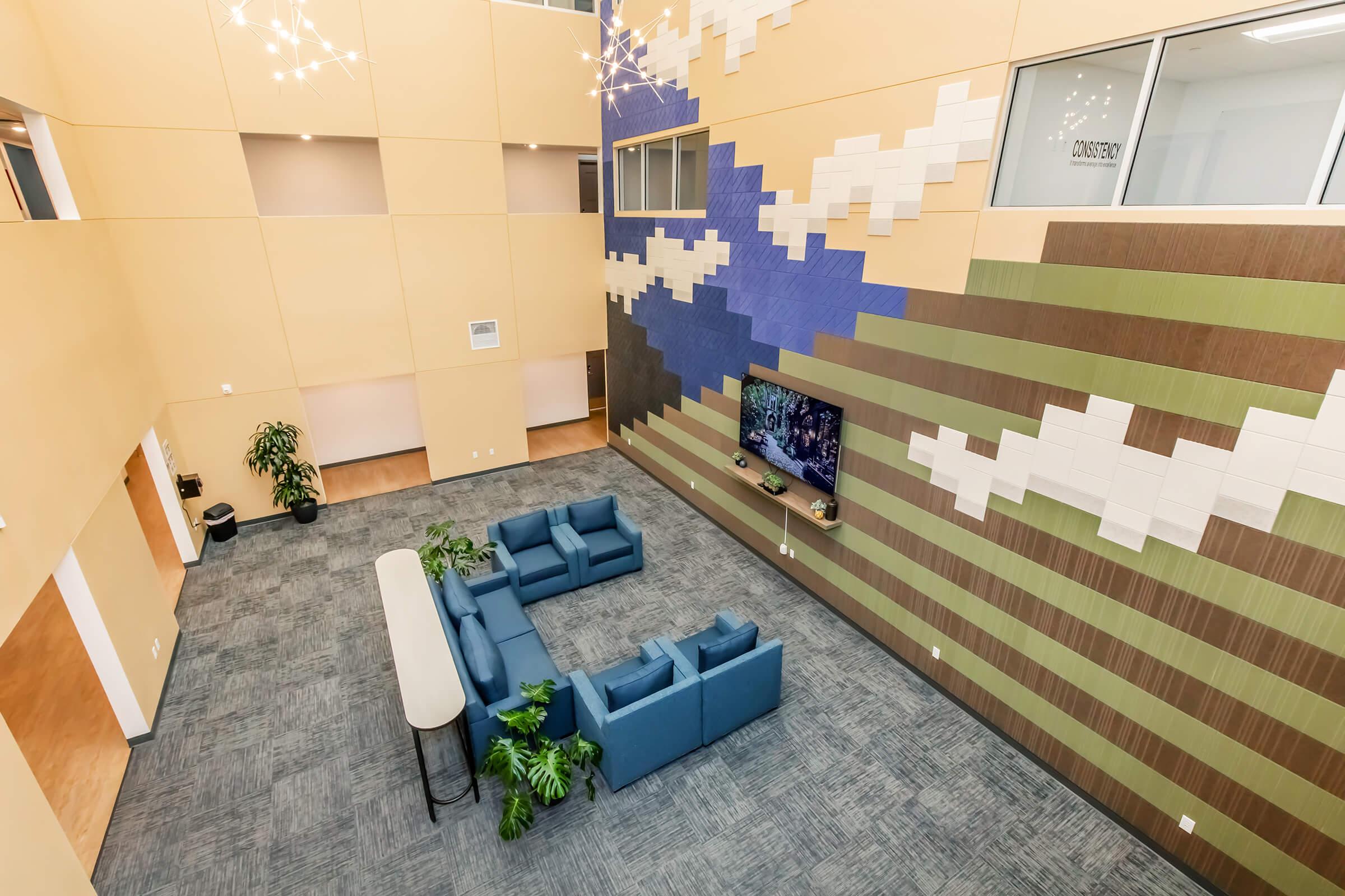 A modern interior space featuring a lounge area with blue couches and a long table. The walls are decorated with colorful geometric patterns and a large screen displaying visuals. Plants are placed around the seating area, and large windows allow natural light to fill the room.