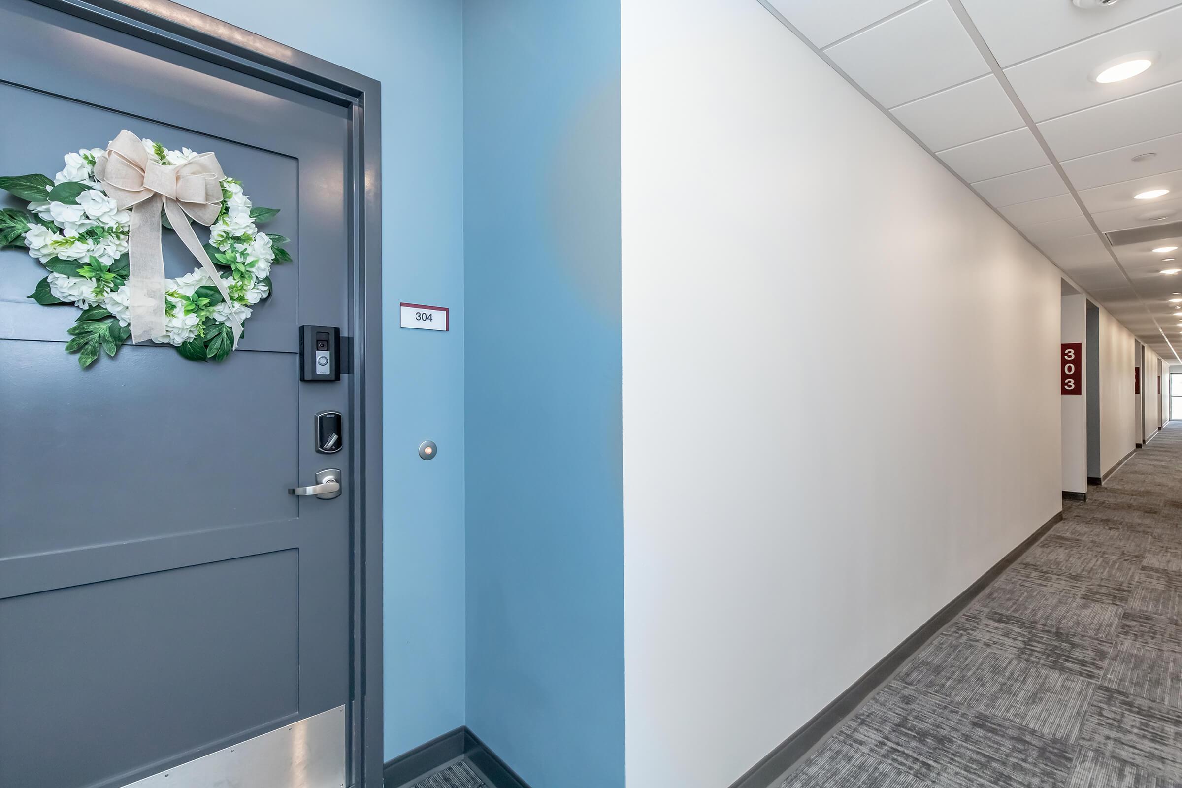 A hallway with light blue walls and gray carpeting. The focus is on the door to room 304, adorned with a floral wreath, beside a wall with a room number sign. A glimpse of the adjacent door, labeled 303, is visible along the corridor, which is brightly lit and features overhead lights.