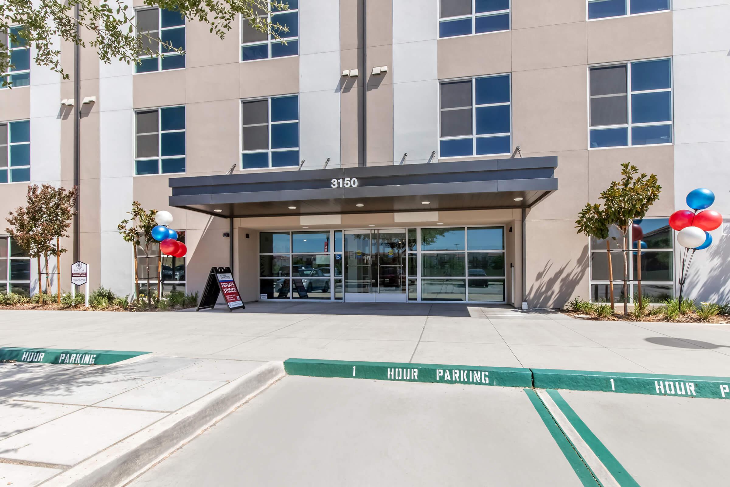 Exterior view of a modern building with large glass entrance doors. The entrance features a canopy and is flanked by decorative trees. Red and blue balloons are tied to pillars on either side. A sign by the entrance advertises parking details, and the pavement is marked for one-hour parking.