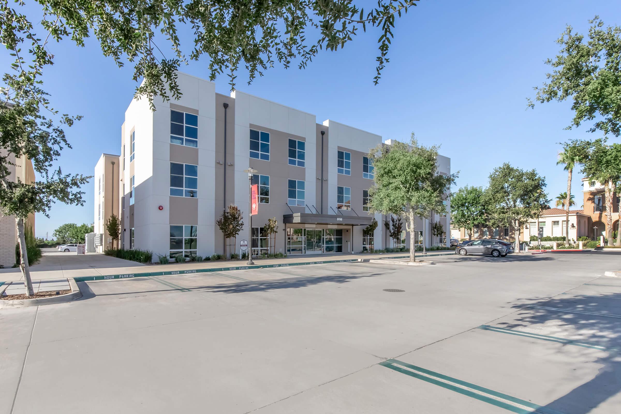 Modern multi-story building with large windows and a landscaped entrance, set against a clear blue sky. The surrounding area features trees and a spacious parking lot.