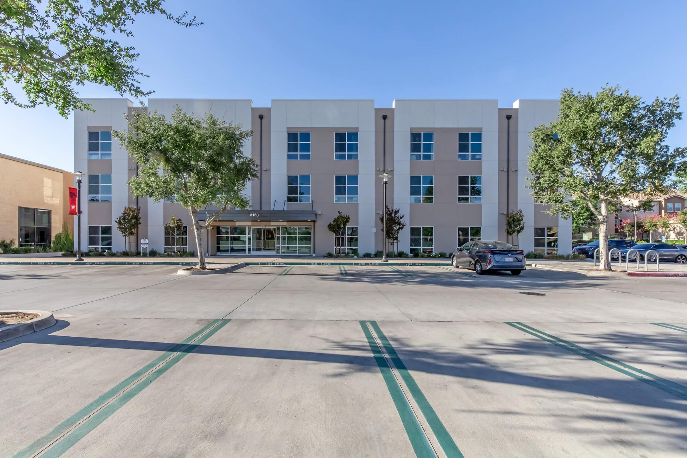 A modern two-story hotel building featuring a light-colored facade and large windows. The front entrance is framed by trees and greenery. A spacious parking lot is visible in the foreground with marked parking spaces. Clear blue skies complete the scene, giving a welcoming appearance.