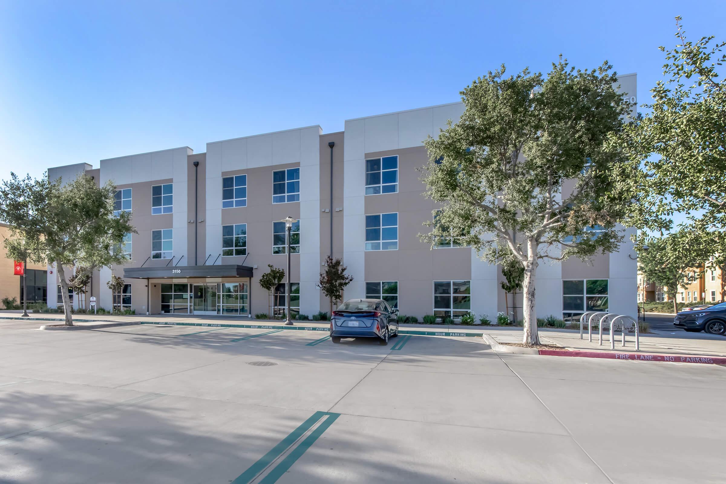 Modern three-story commercial building with a light-colored exterior and large windows. The front entrance features a canopy. Surrounding the building are several trees and well-maintained landscaping. A parking lot in the foreground includes a gray car and bike racks. The sky is clear and blue.