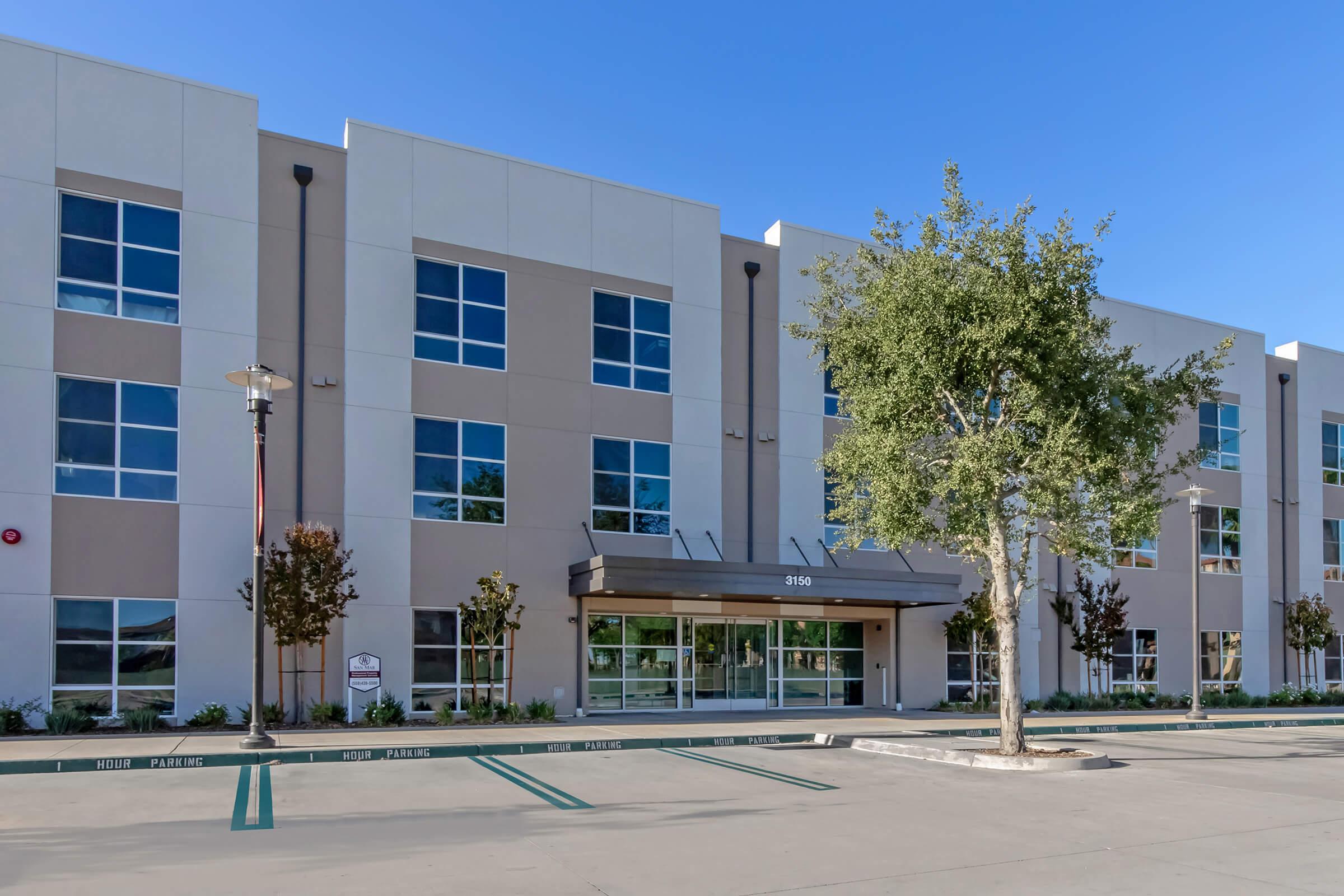 Modern building exterior with a flat facade, large windows, and a canopy entrance. Landscaping includes trees and decorative plants. The parking lot in front has marked spaces, and the sky is clear and blue.