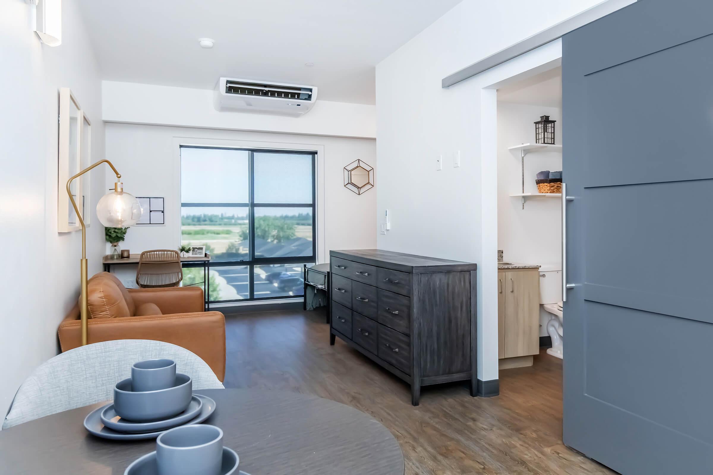 A modern, minimalistic living space featuring a cozy brown armchair, a wooden dresser, and a round dining table set with dishes. A large window offers a view of the outdoors, while a door leads to a bathroom area. The walls are light-colored, and stylish decor items are present throughout the room.
