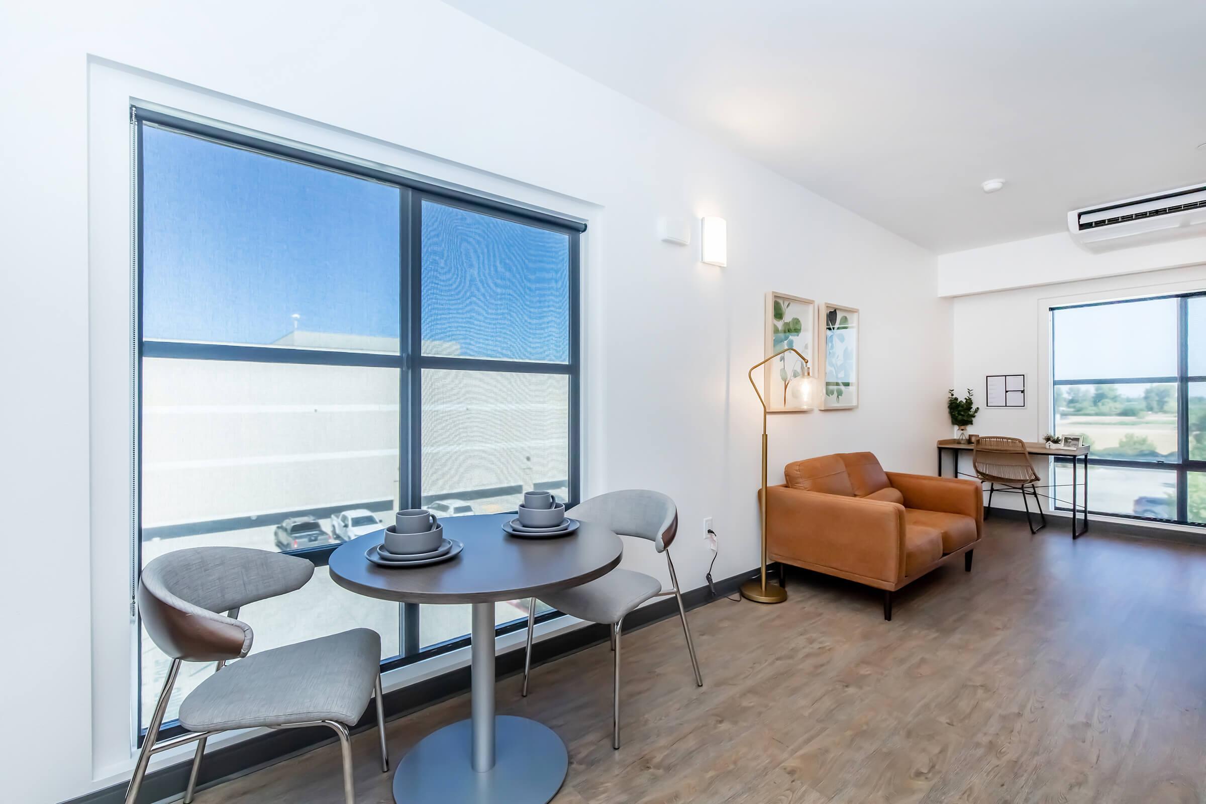 Modern living space featuring a cozy brown leather sofa, a small round table set for two with gray chairs, and large windows allowing natural light. A stylish floor lamp and a desk area are visible, creating a warm and inviting atmosphere.
