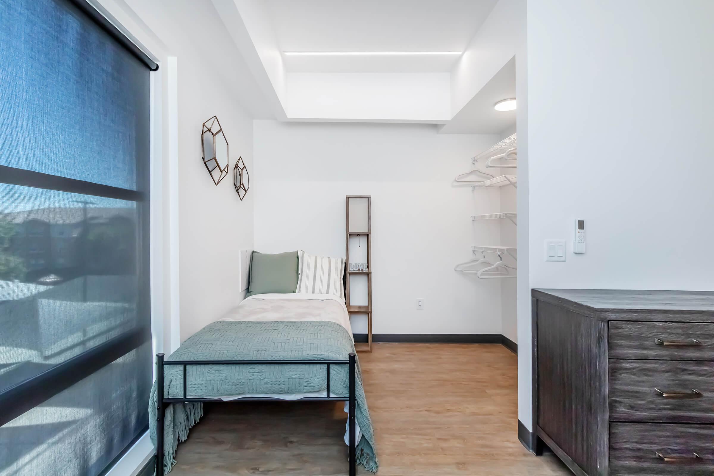 A small, minimalistic bedroom featuring a single bed with a green throw blanket, a wall-mounted shelving unit, and two decorative wall mirrors. Natural light filters through a window with a blue blind, while a wooden dresser is placed against one wall. The floor is a light wood finish.