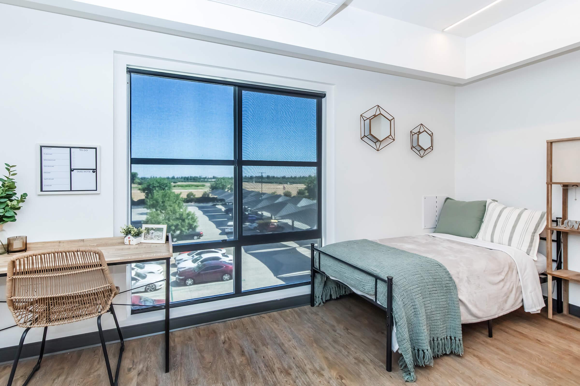 A modern bedroom featuring a bed with a light blanket, a small desk with a decorative plant, and a window offering a view of a parking lot and outdoor scenery. The room has stylish wall decor and a light, inviting atmosphere.
