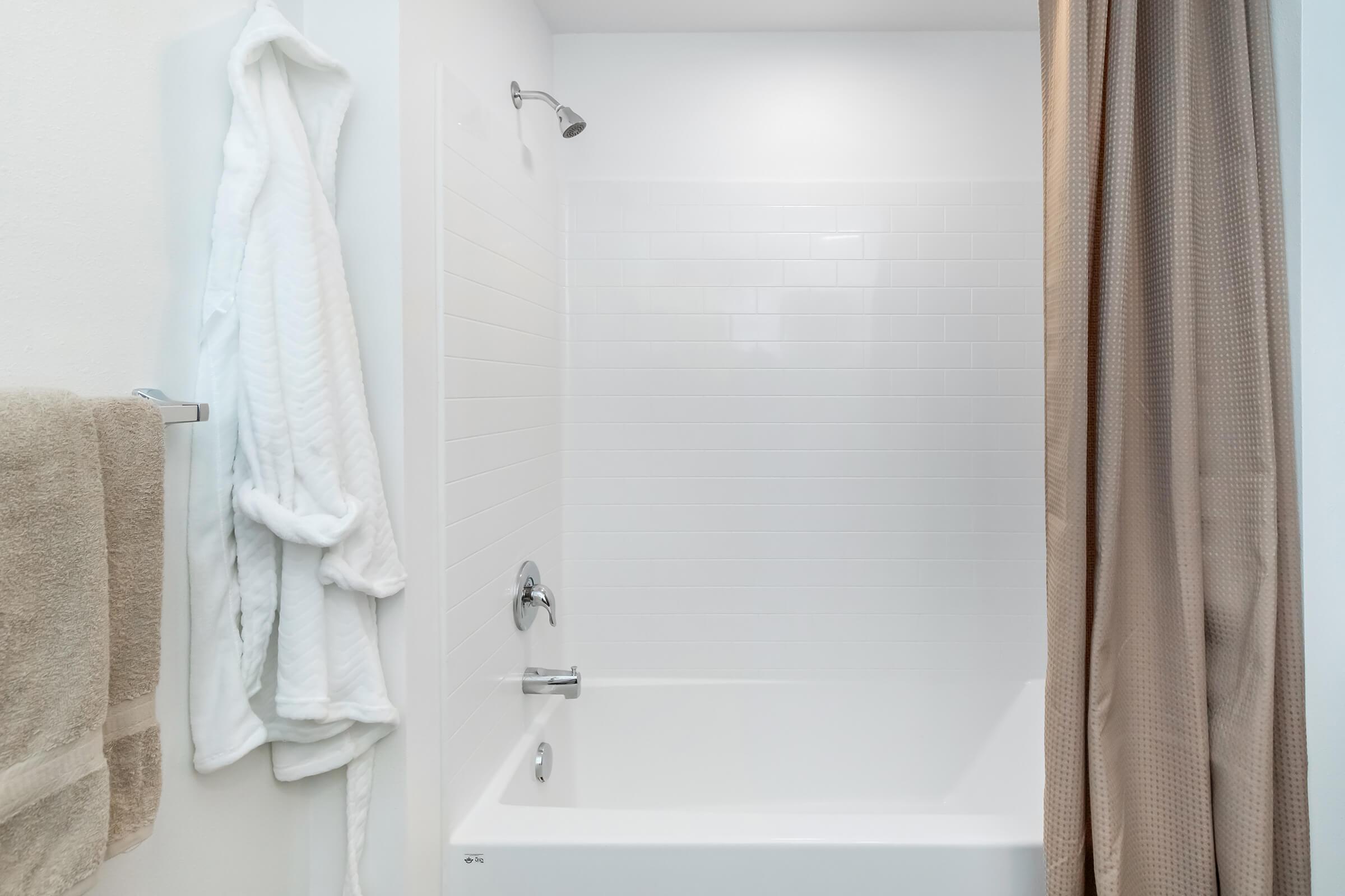 A clean, minimalistic bathroom featuring a white bathtub, a shower with a chrome faucet, and a beige shower curtain. A fluffy white bathrobe hangs on a towel rack beside a cream-colored towel, enhancing the cozy atmosphere. The walls are white and accented with tile.